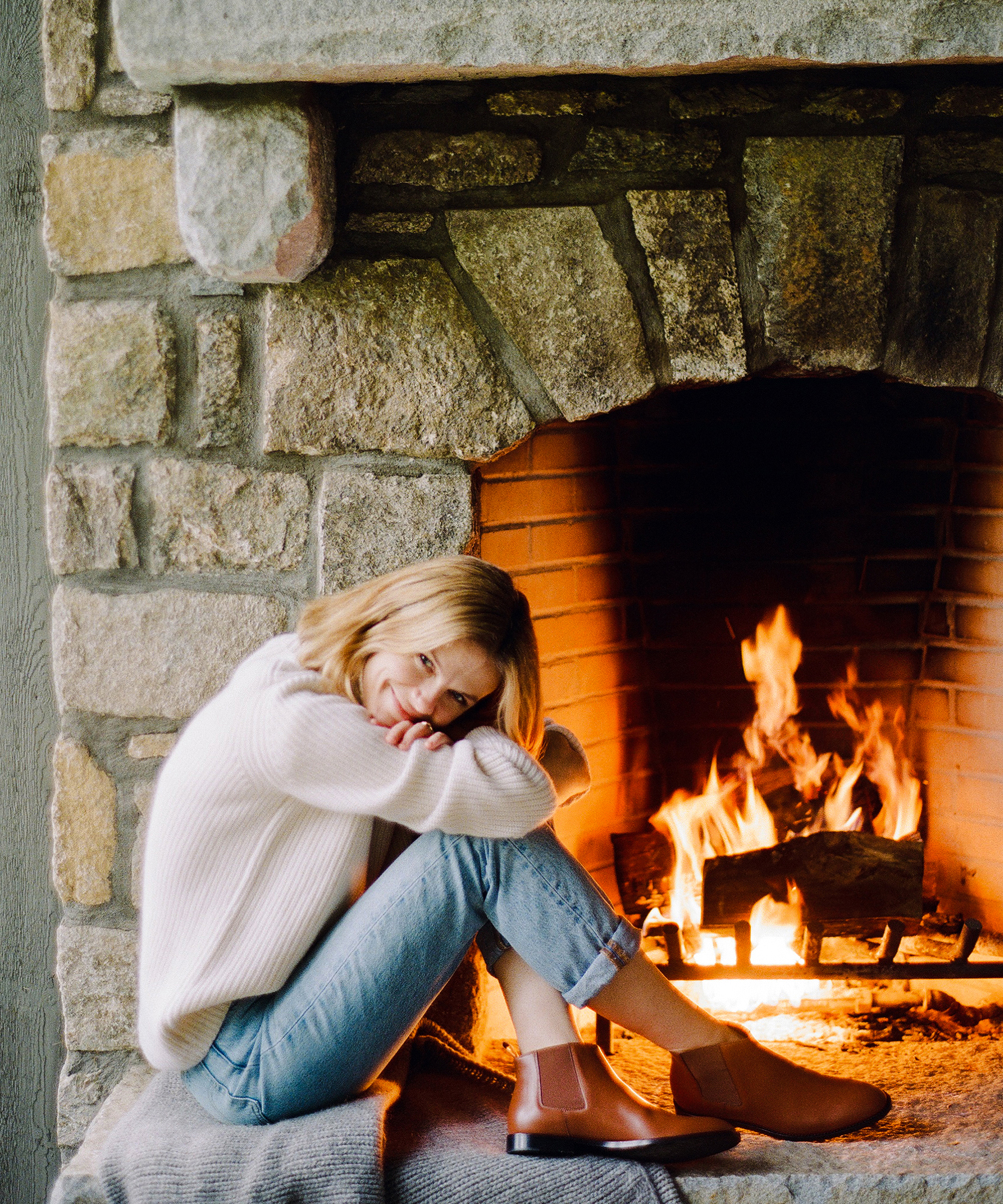 A woman, resembling Brooklyn Decker, in a white sweater and jeans sits on a blanket by a stone fireplace with a warm fire, resting her head on her knees and smiling gently.