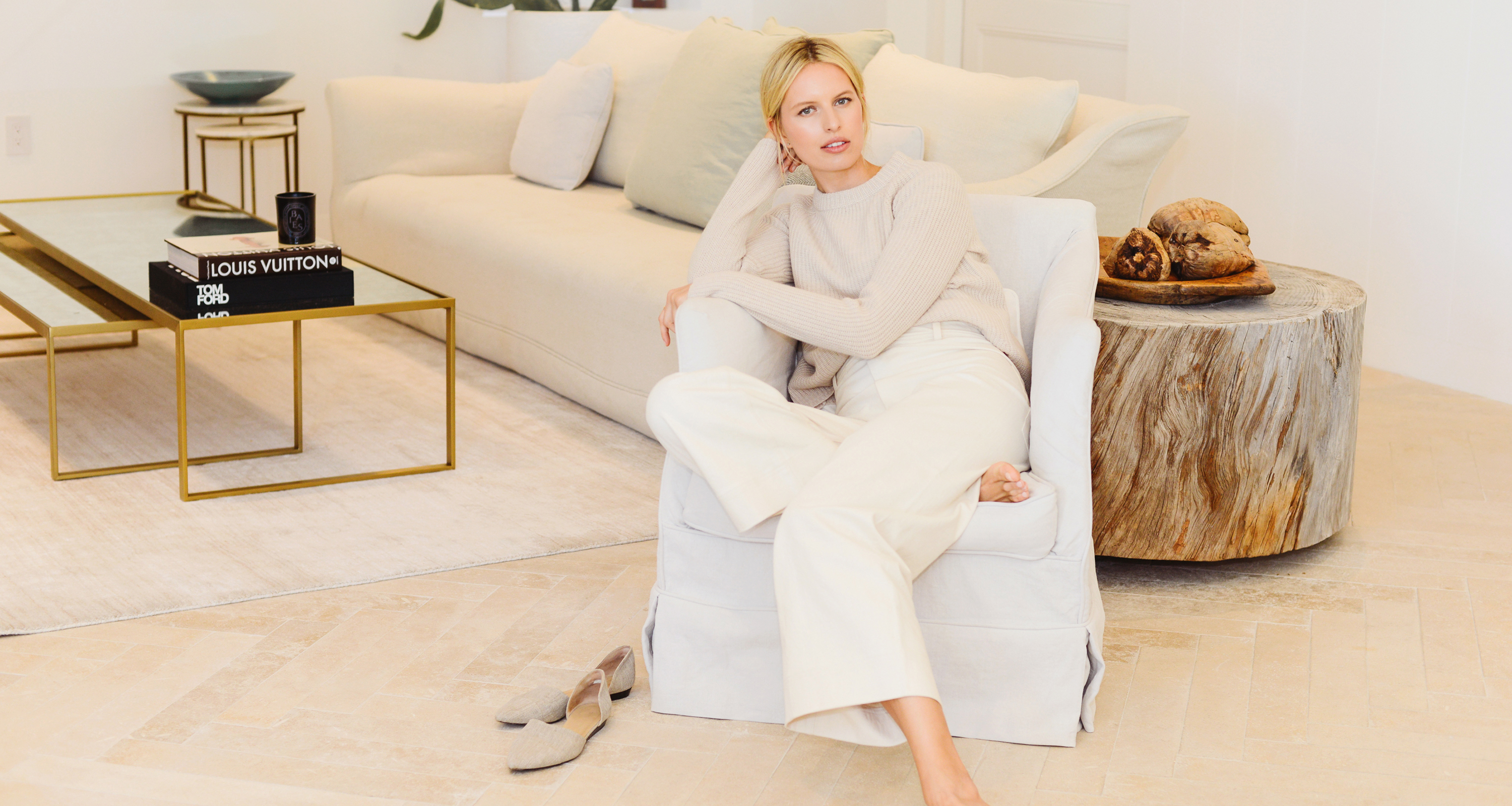 A woman in a beige sweater and white pants sits barefoot in a white armchair, with her shoes on the floor nearby. She rests her head on one hand, surrounded by modern, neutral-toned decor in a stylish living room.