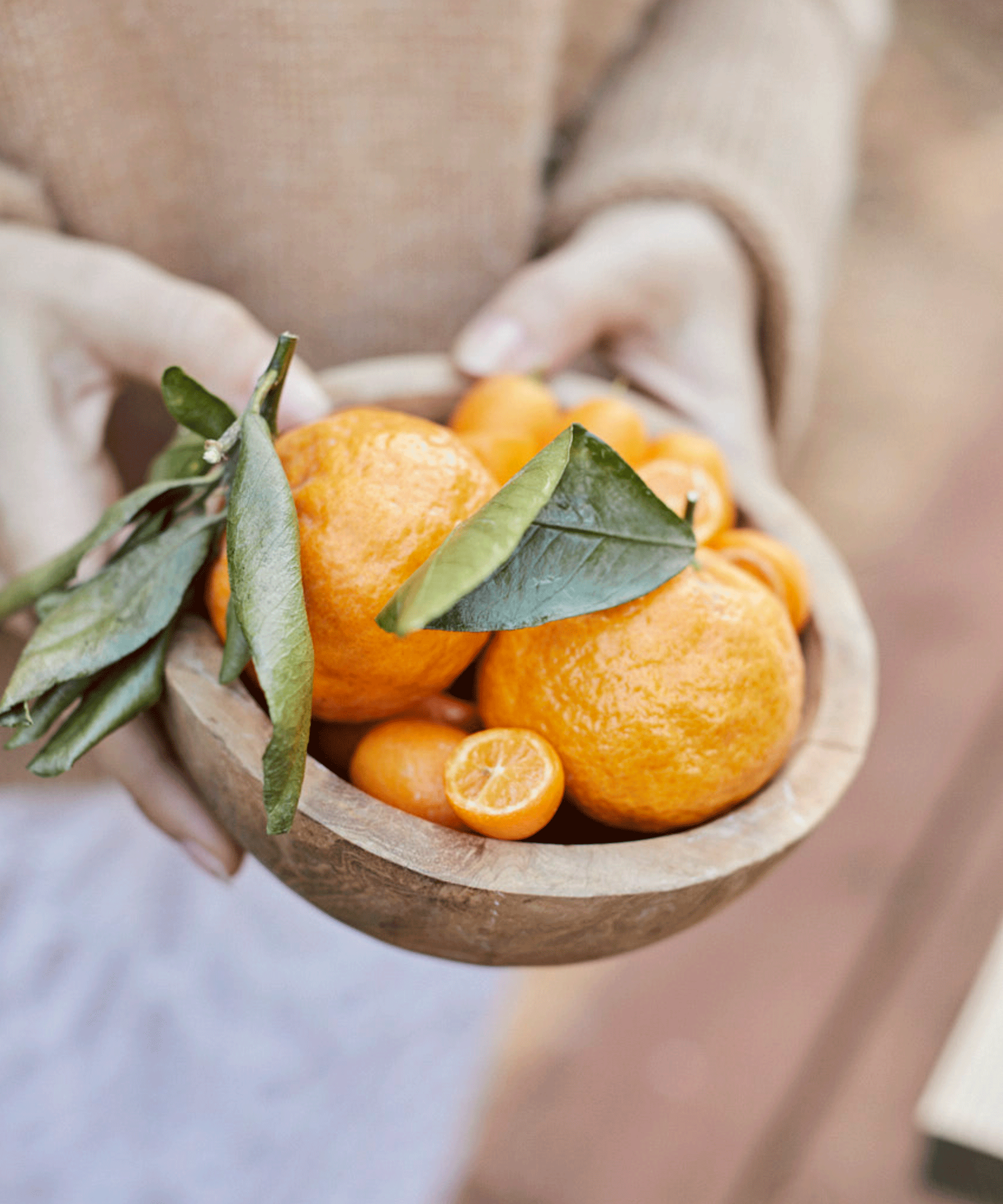 A person in a beige sweater holds a wooden bowl filled with fresh tangerines, some with green leaves still attached.