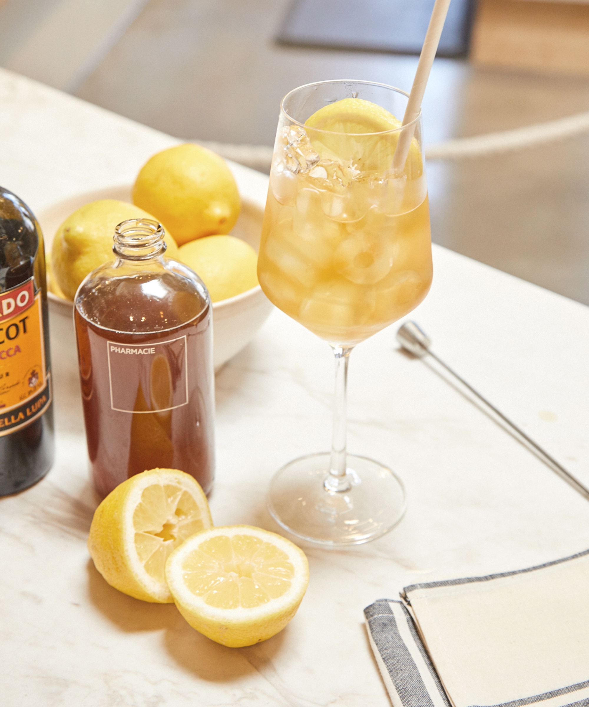 A glass of iced tea garnished with a lemon slice and straw sits on a marble counter, surrounded by whole and cut lemons, a bottle of dark liqueur, Aperol Spritz syrup, and a folded napkin.