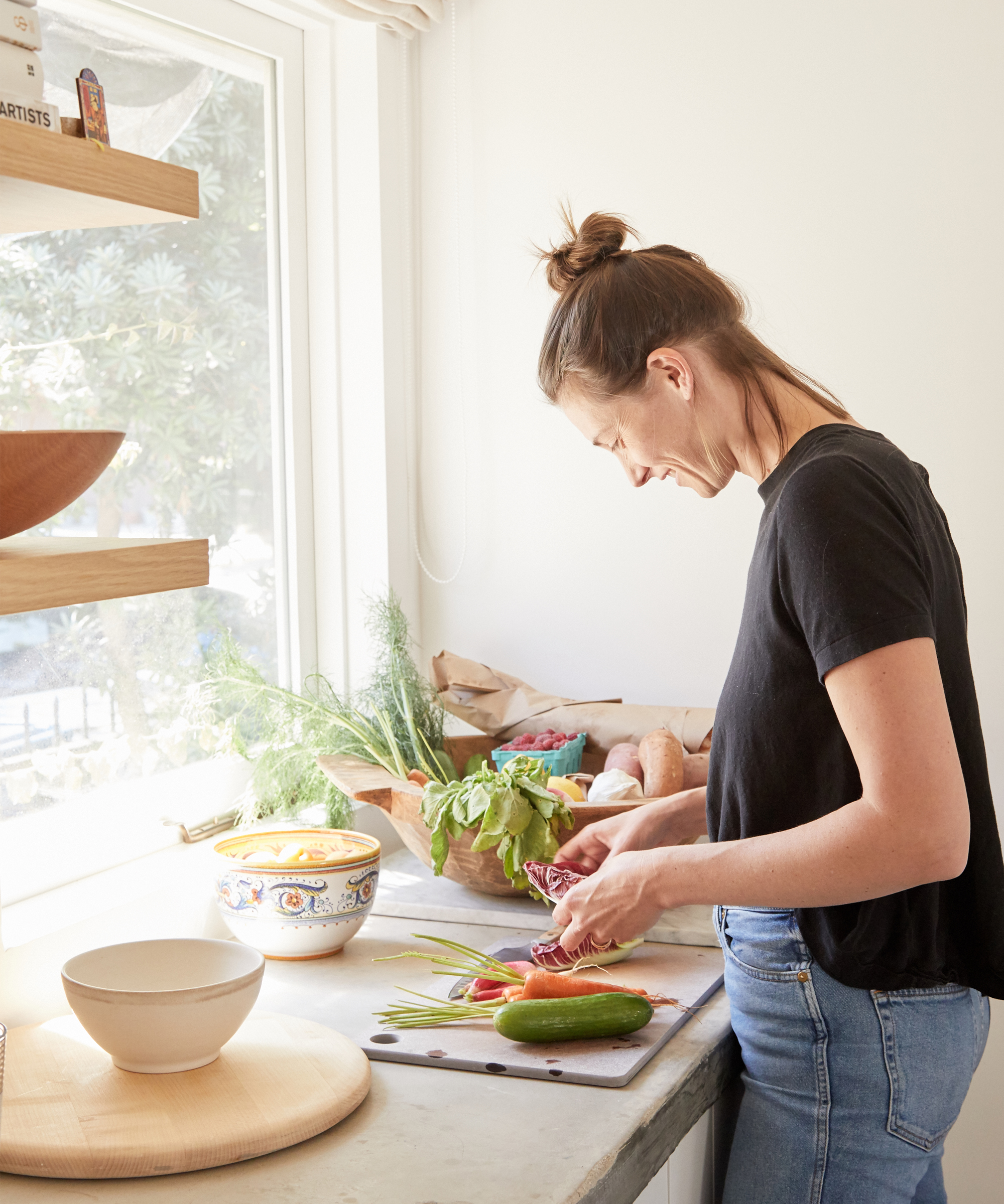 A woman stands at a kitchen counter near a bright window, chopping vegetables including cucumber and red cabbage. Fresh herbs and produce fill the space, inspiring light, airy Summer Dinner Recipes in this inviting atmosphere.