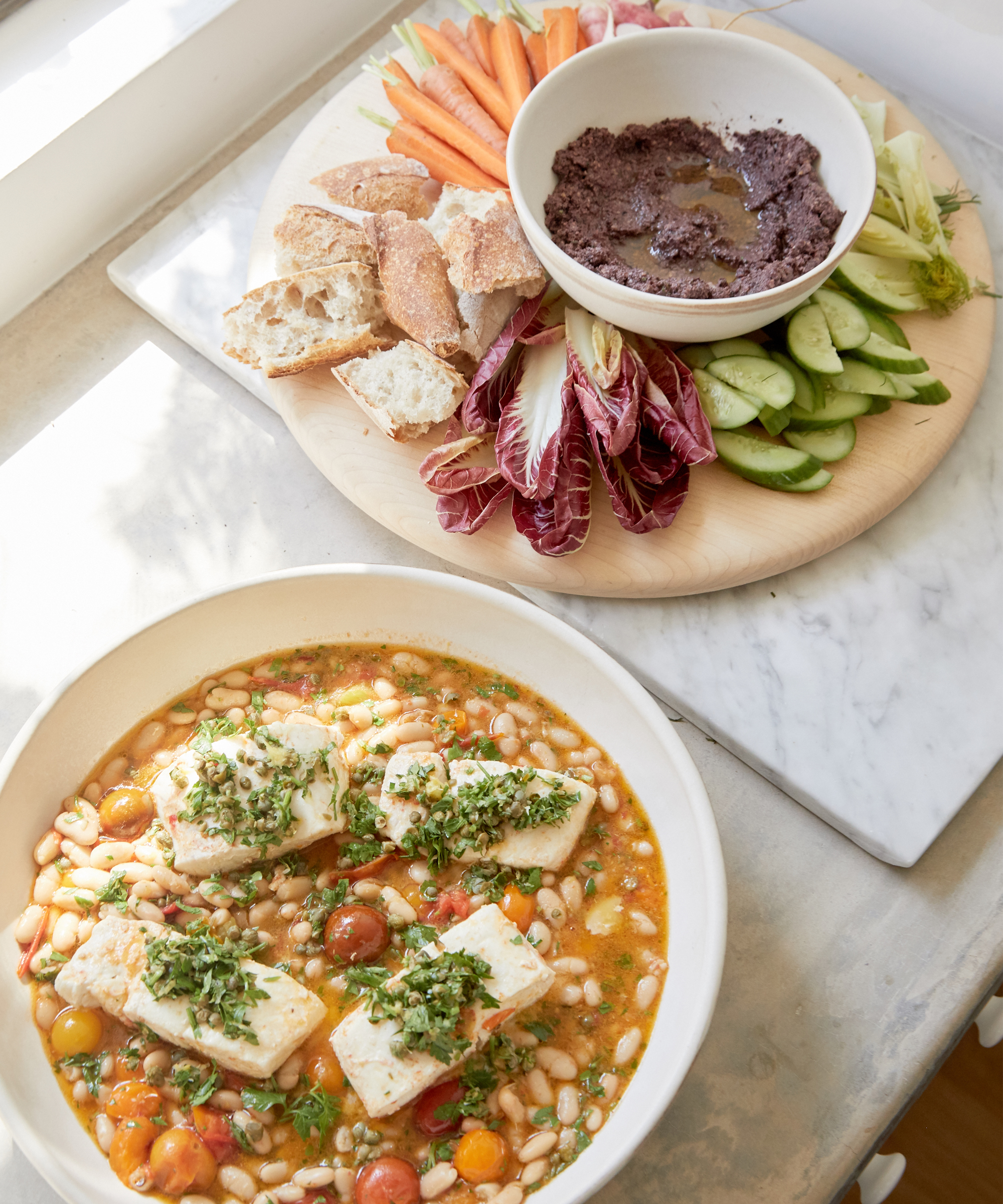 A perfect pick for Summer Dinner Recipes: a table with a bowl of fish topped with herbs over white beans and cherry tomatoes, accompanied by a platter of sliced bread, veggie sticks, and a rich, dark dip.