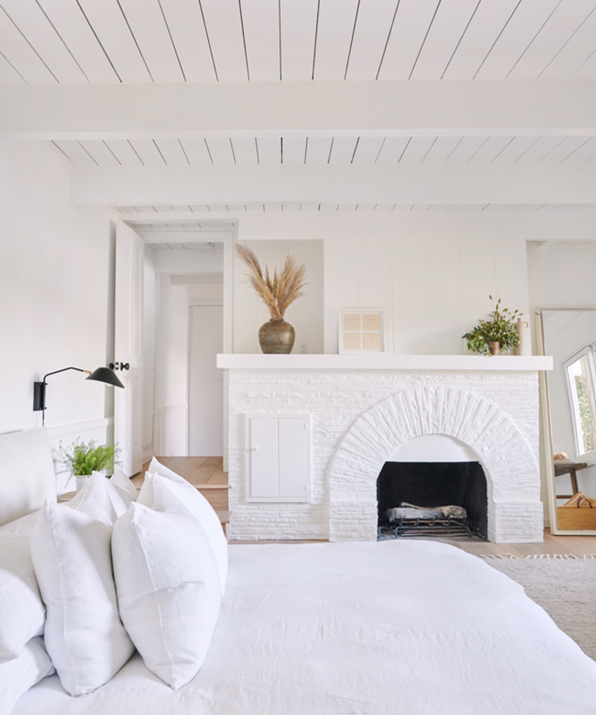 Bright, airy lake house bedroom with white walls, ceiling, bedding, and a white-painted brick fireplace. Decorative vases with pampas grass and greenery sit on the mantel, creating a calm, minimalist aesthetic.