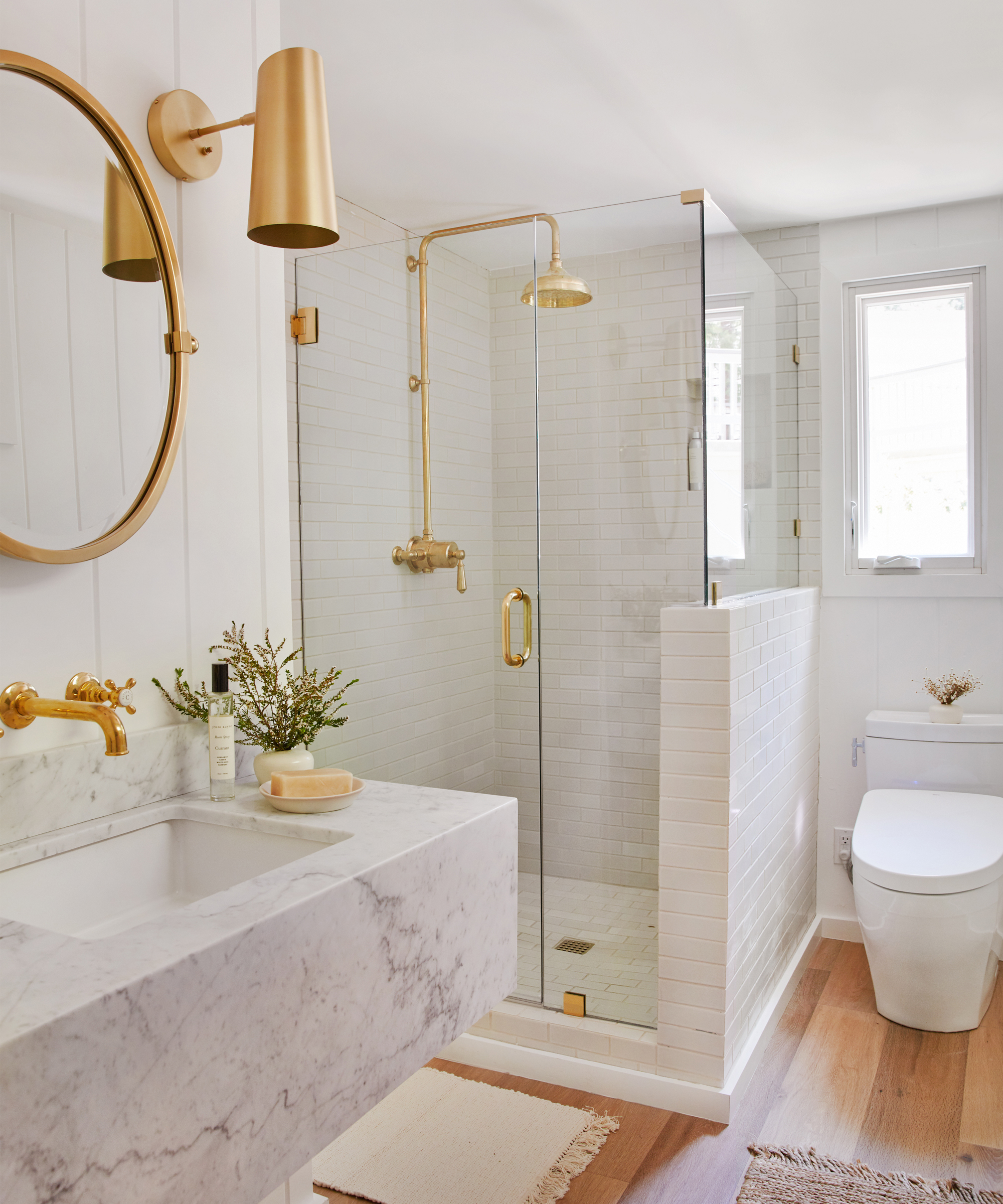 A modern lake house bathroom with a marble sink, gold fixtures, a glass-enclosed shower, white tiled walls, a round mirror, wall sconce, window, toilet, and wooden flooring.
