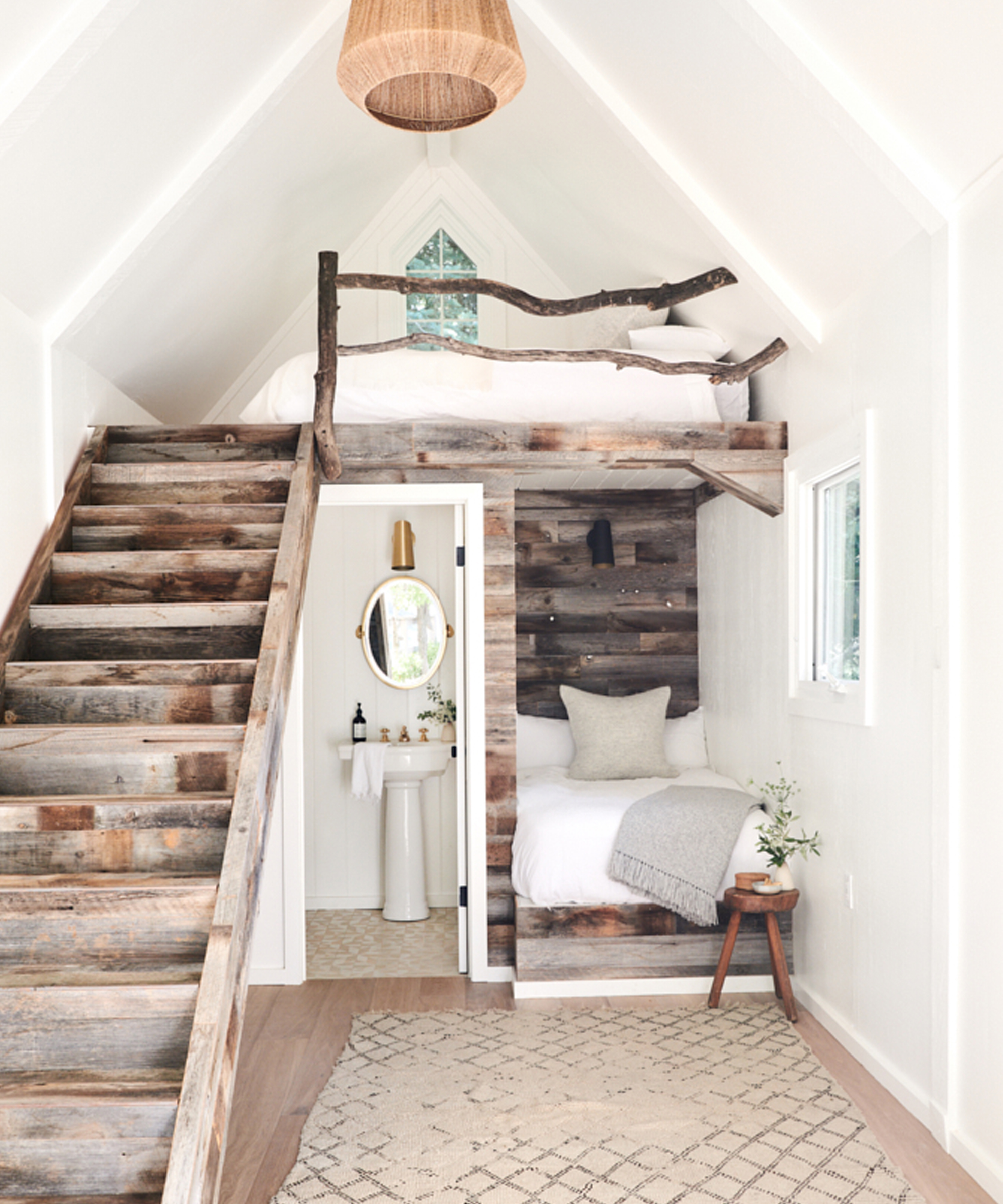 A cozy lake house tiny interior features rustic wooden stairs leading to a loft bed with a branch railing, a lower bed nook, and a small bathroom with a round mirror. Neutral decor and soft lighting create a warm atmosphere.