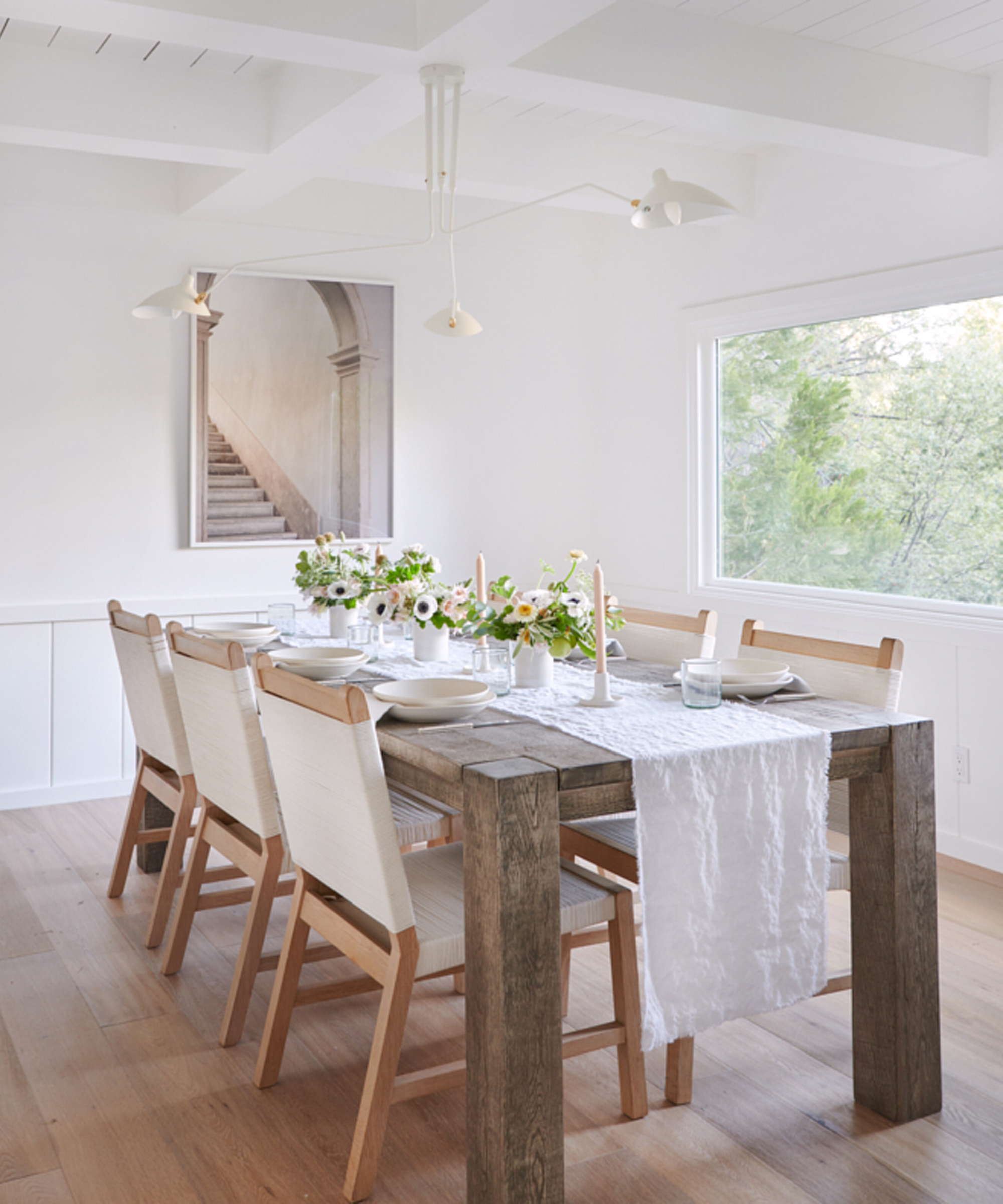 A bright dining room styled for a lake house, featuring a rustic wooden table, six light wood chairs, a white table runner, floral centerpieces, candles, white walls, a large window, and a framed stairway photograph on the wall.