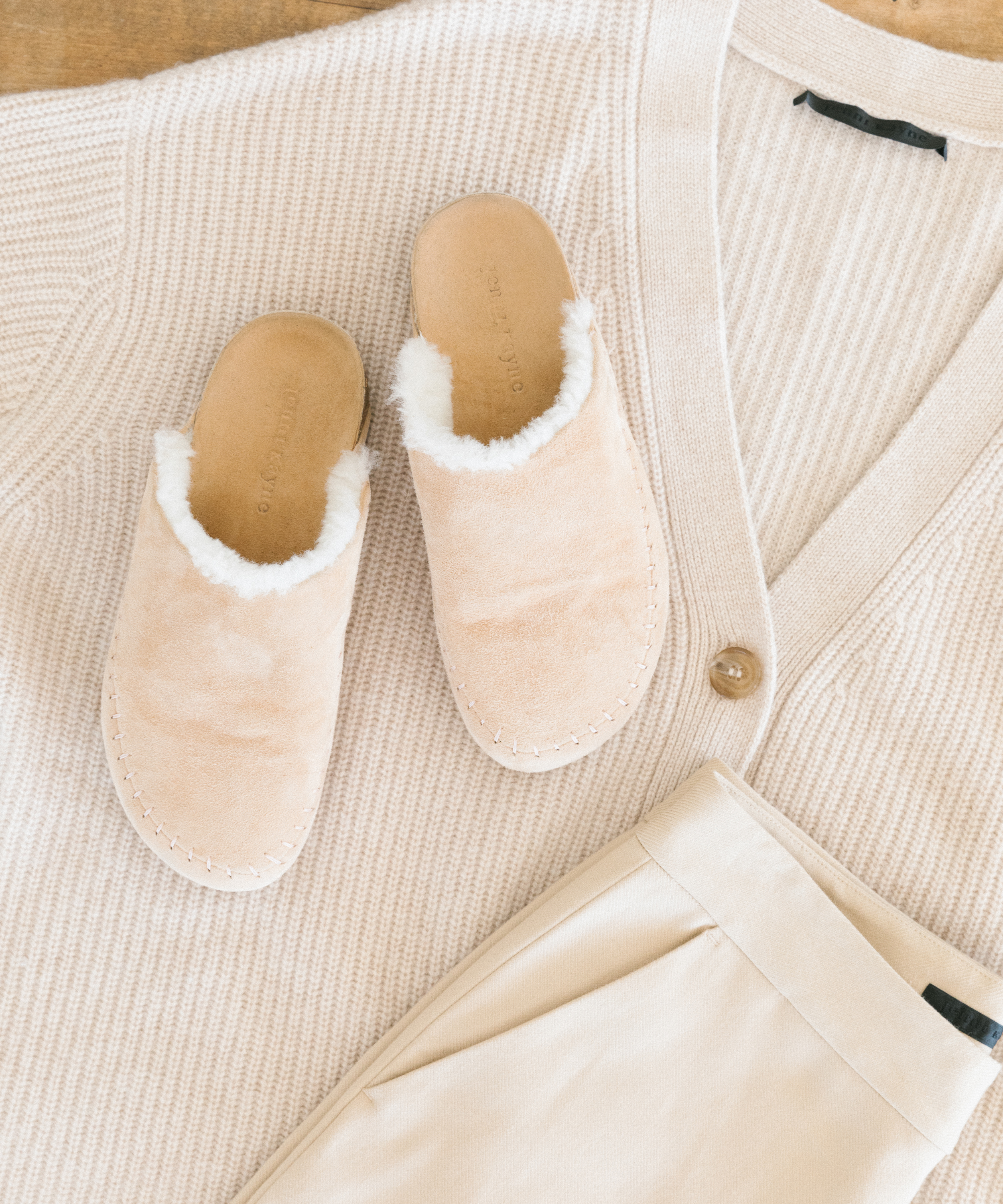 A pair of beige, fuzzy slippers—styled like the best moccasins—rests on a cream-colored knit cardigan and light beige pants, all laid out in a coordinated, neutral-toned arrangement.