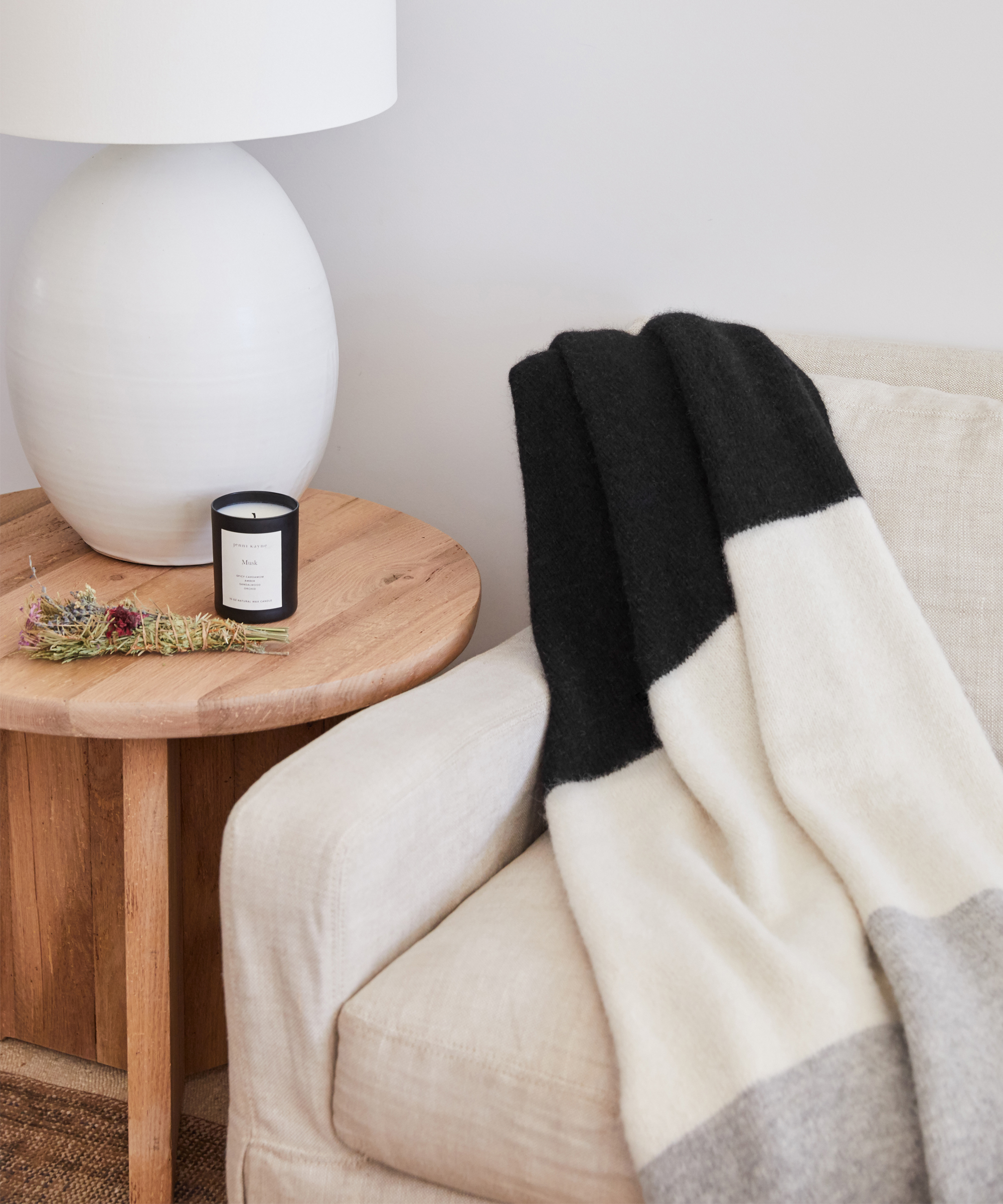 A cozy living room scene perfect for holiday hosting, featuring a beige armchair draped with a black, white, and gray color-blocked blanket beside a round wooden side table holding a white lamp, glass candle, and small dried flower arrangement.