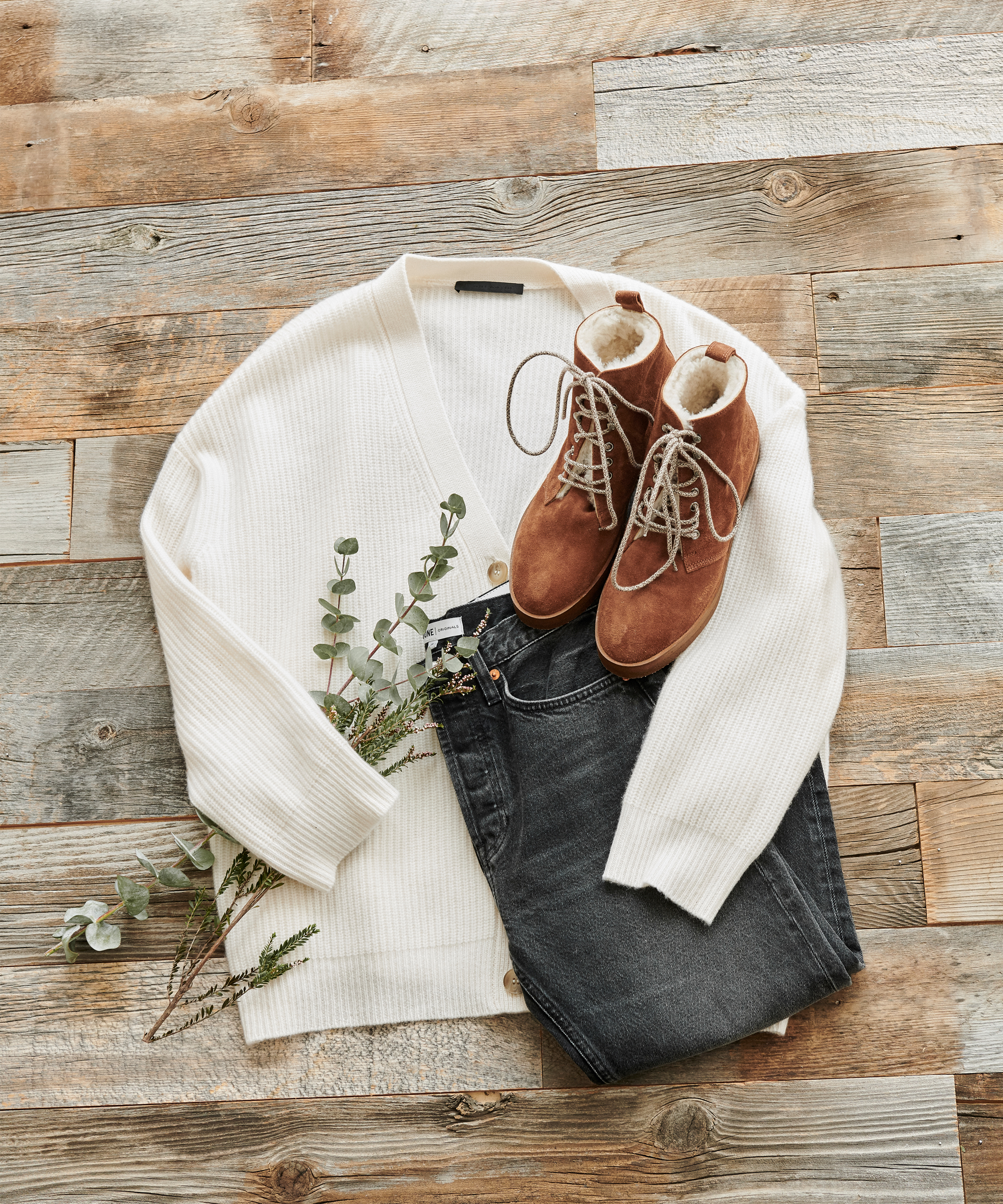 A cozy 2020 style outfit laid on a wooden surface: a white knit cardigan, dark jeans, brown lace-up boots with soft lining, and a sprig of eucalyptus for decoration.