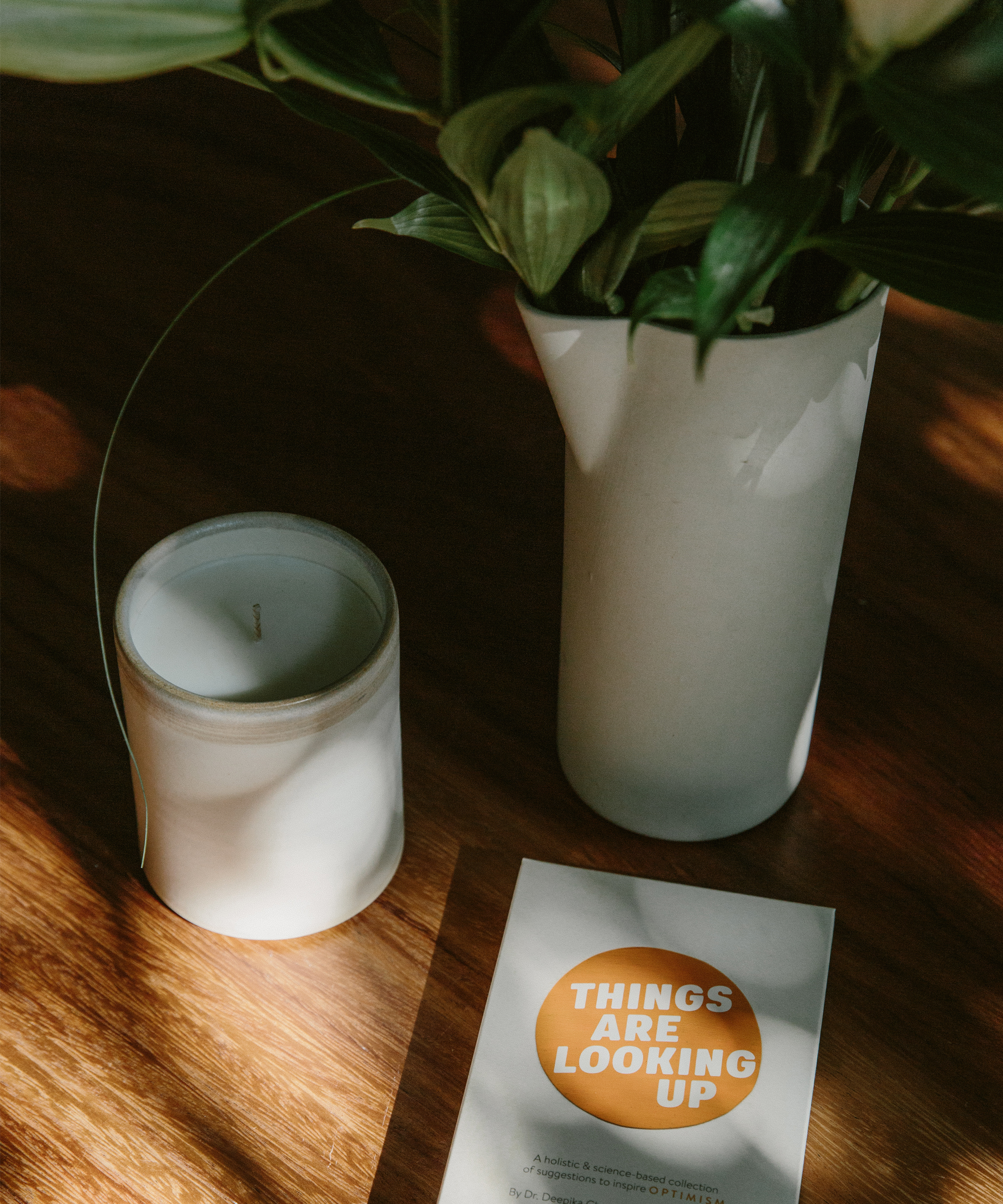 A white vase with green leaves, a white candle, and a card that says “THINGS ARE LOOKING UP” in orange are arranged on a wooden surface in natural sunlight, inspired by optimism doctor Deepika Chopra’s positive outlook.