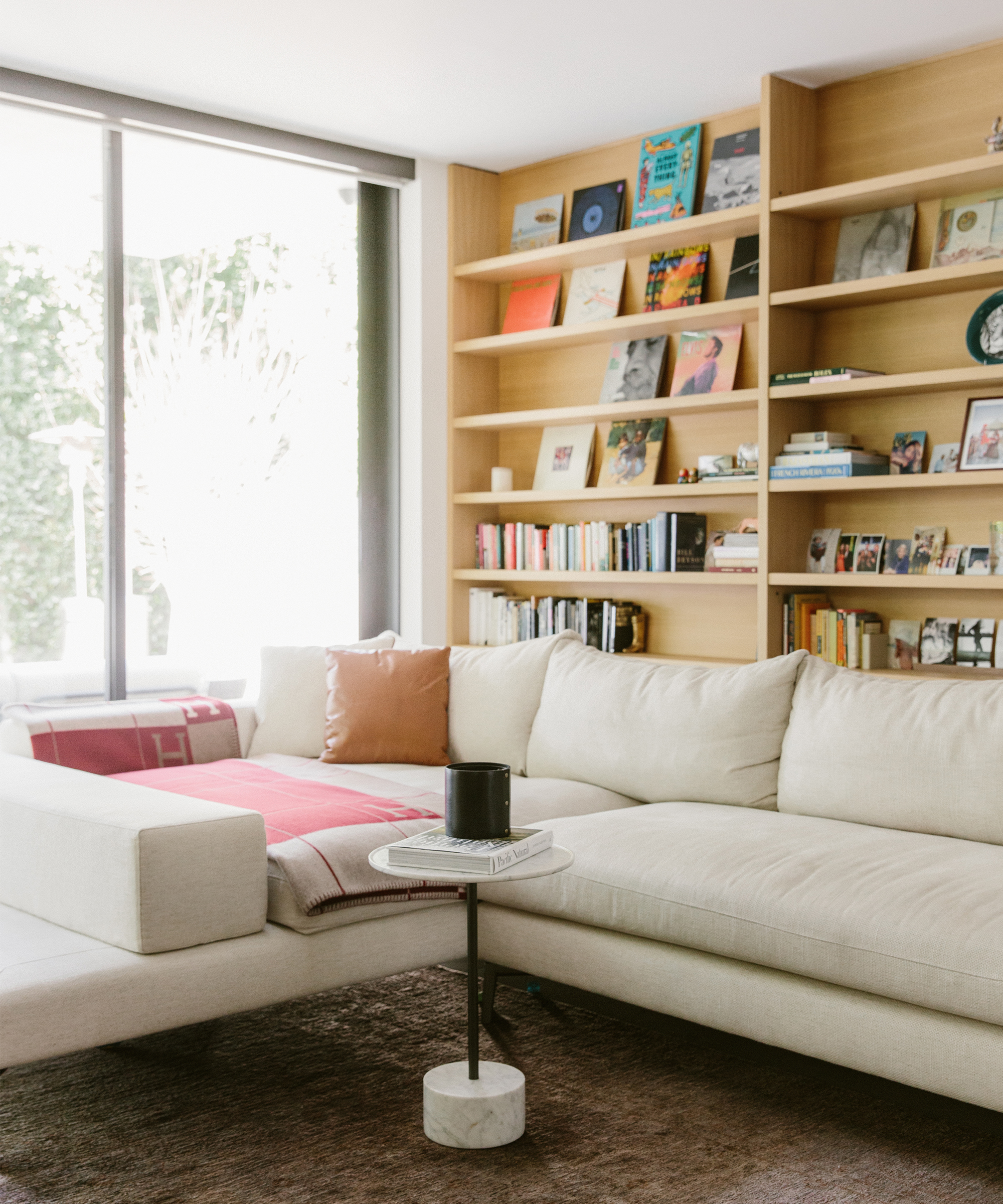 A cozy living room with a large cream sectional sofa, a red blanket, a brown pillow, and built-in wooden bookshelves filled with books and decor by a large window—perfect for relaxing or reading works by optimism doctor Deepika Chopra.