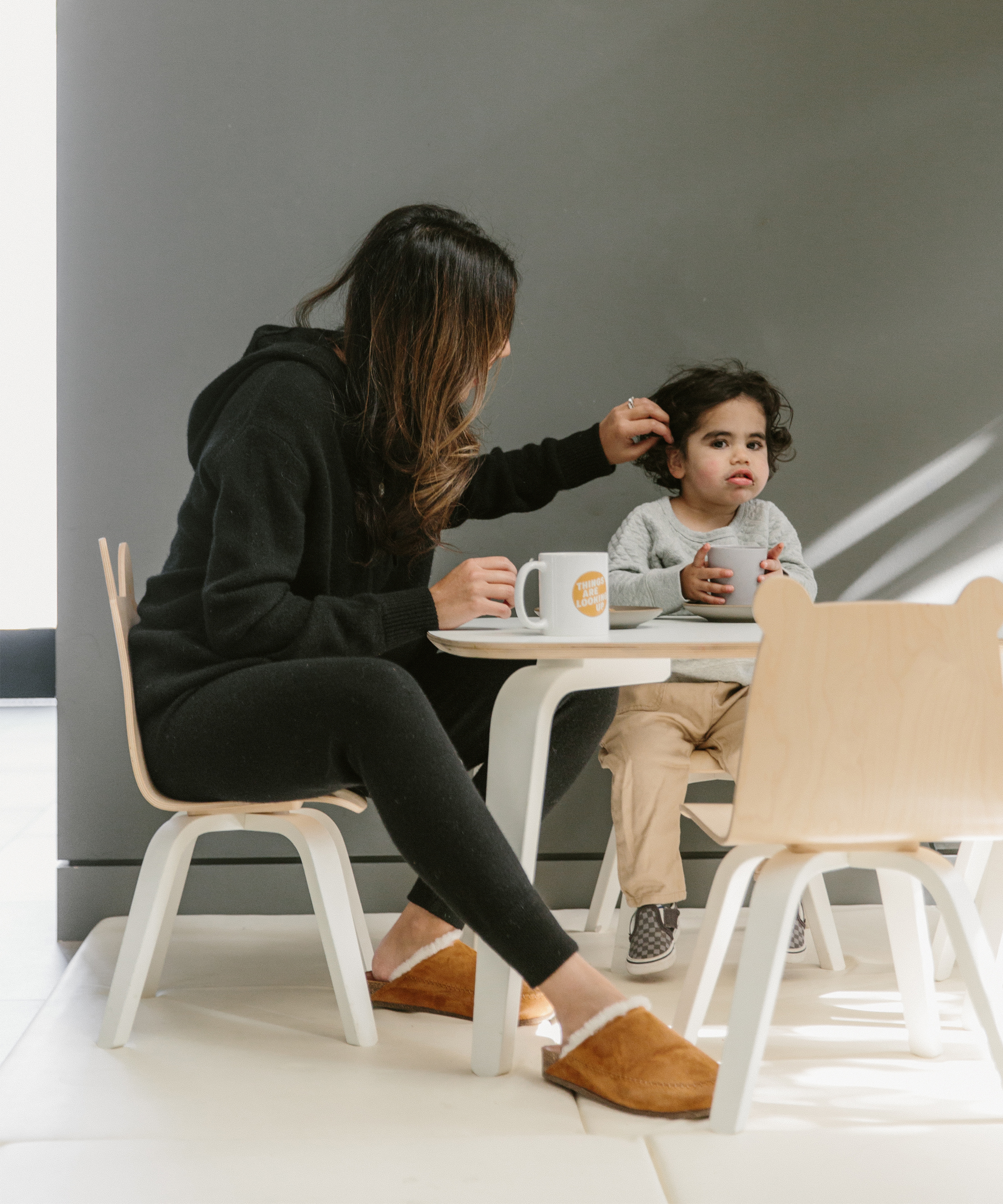 A woman, embodying optimism like doctor Deepika Chopra, sits at a child-sized table with a young child, gently fixing the childs hair. The child looks pouty while the woman holds a mug and offers a comforting glance.
