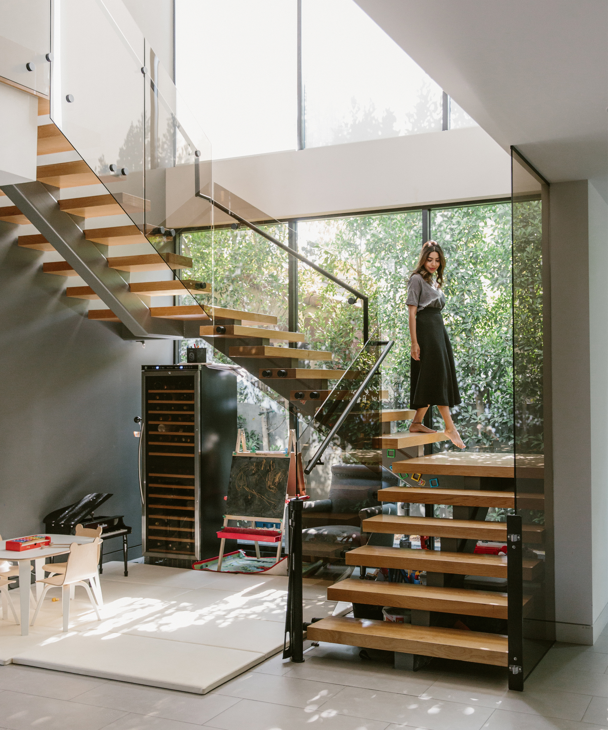 A woman stands on a modern wooden staircase with glass railings in a bright, spacious room with large windows overlooking greenery—a scene radiating optimism reminiscent of Dr. Deepika Chopra’s uplifting style. A children’s play area sits below.