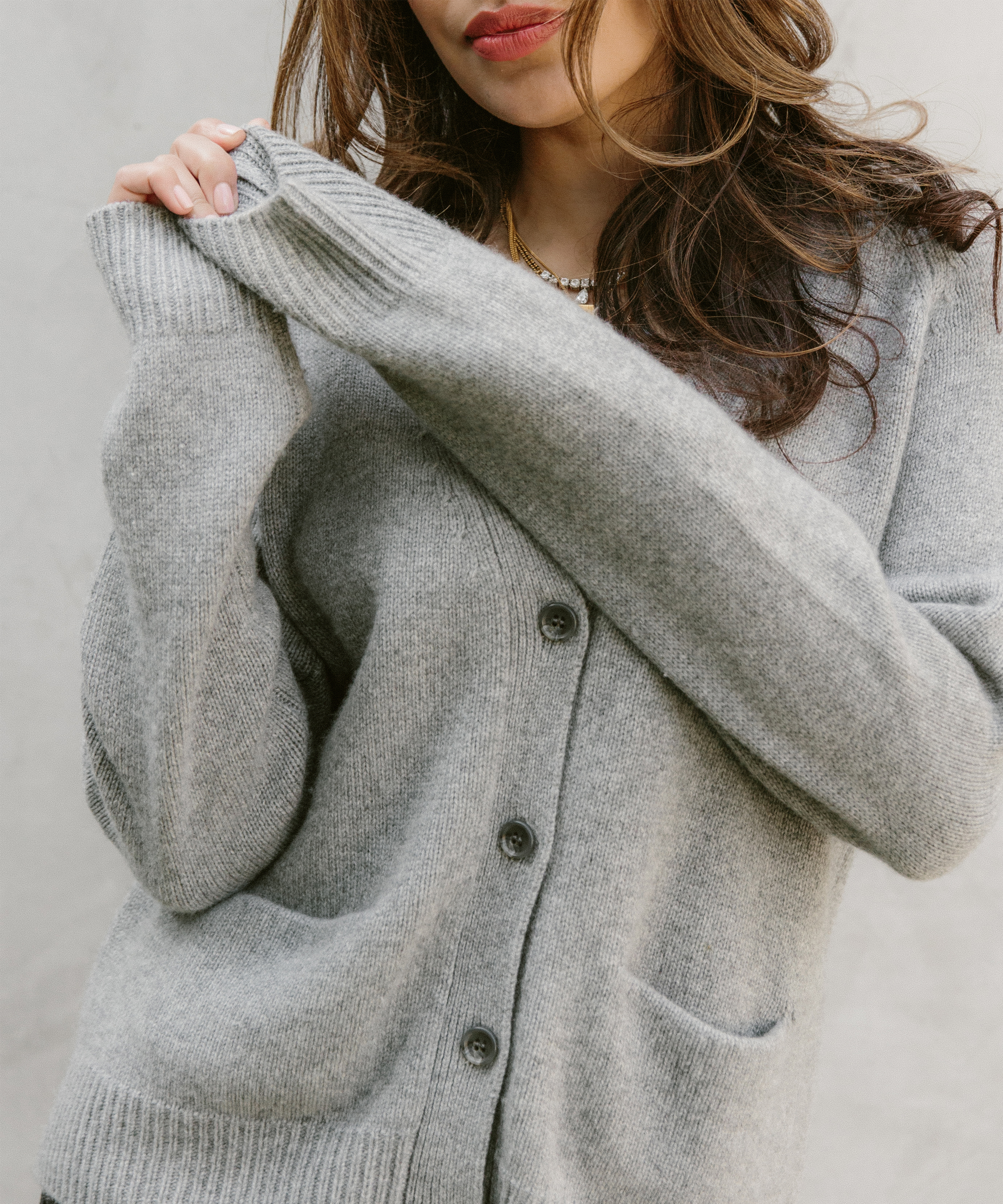 A woman wearing a gray button-up cardigan with front pockets stands against a neutral background. Inspired by optimism doctor Deepika Chopra, she wraps her arms around herself, slightly pulling on her sweater sleeves.