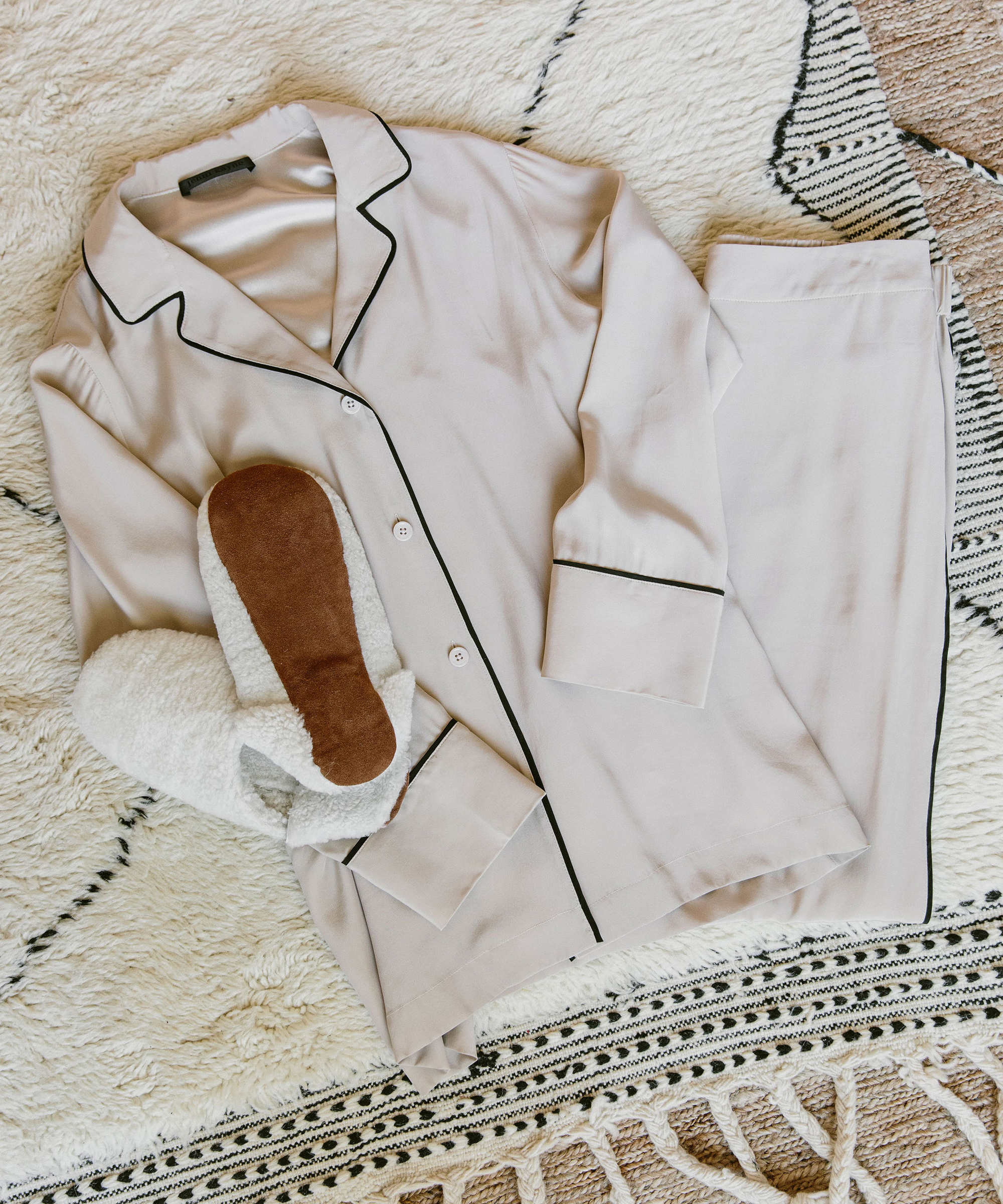 A neatly folded light beige pajama set with black piping, paired with white fuzzy slippers, channels 2020 style as it rests on a cream and black patterned rug.