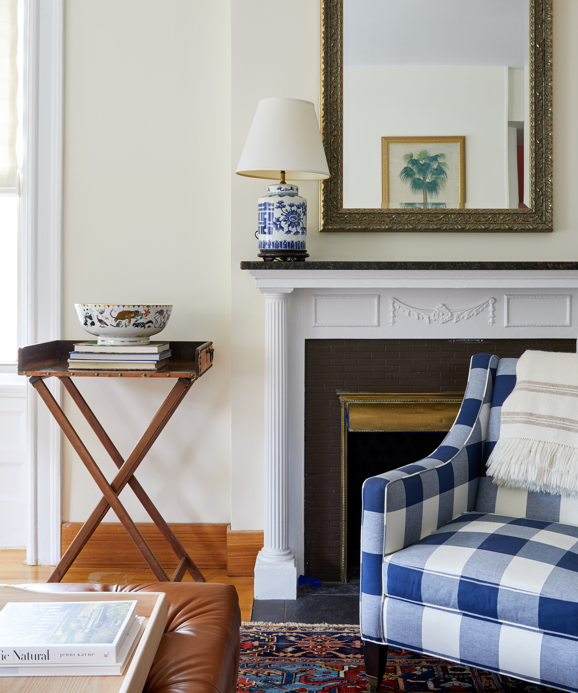 A cozy living room corner from Davina Pike Wovn Home features a blue and white checkered armchair, a cream blanket, a white fireplace with a gold-framed mirror, a lamp, a wooden tray table with books, and a decorative bowl.
