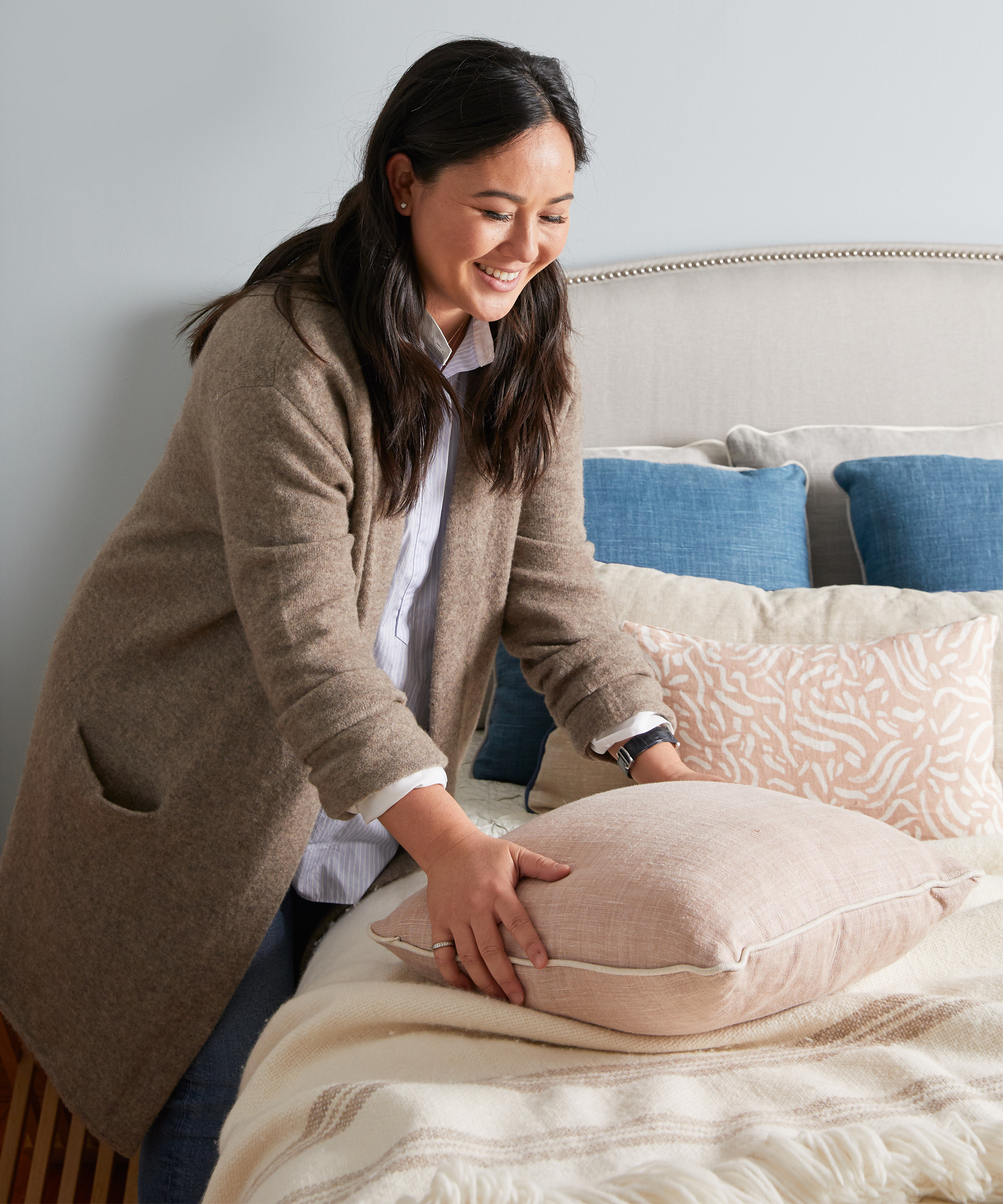 Davin Pike fluffing blush colored pillow on bed wearing oatmeal Jenni Kayne sweater coat.