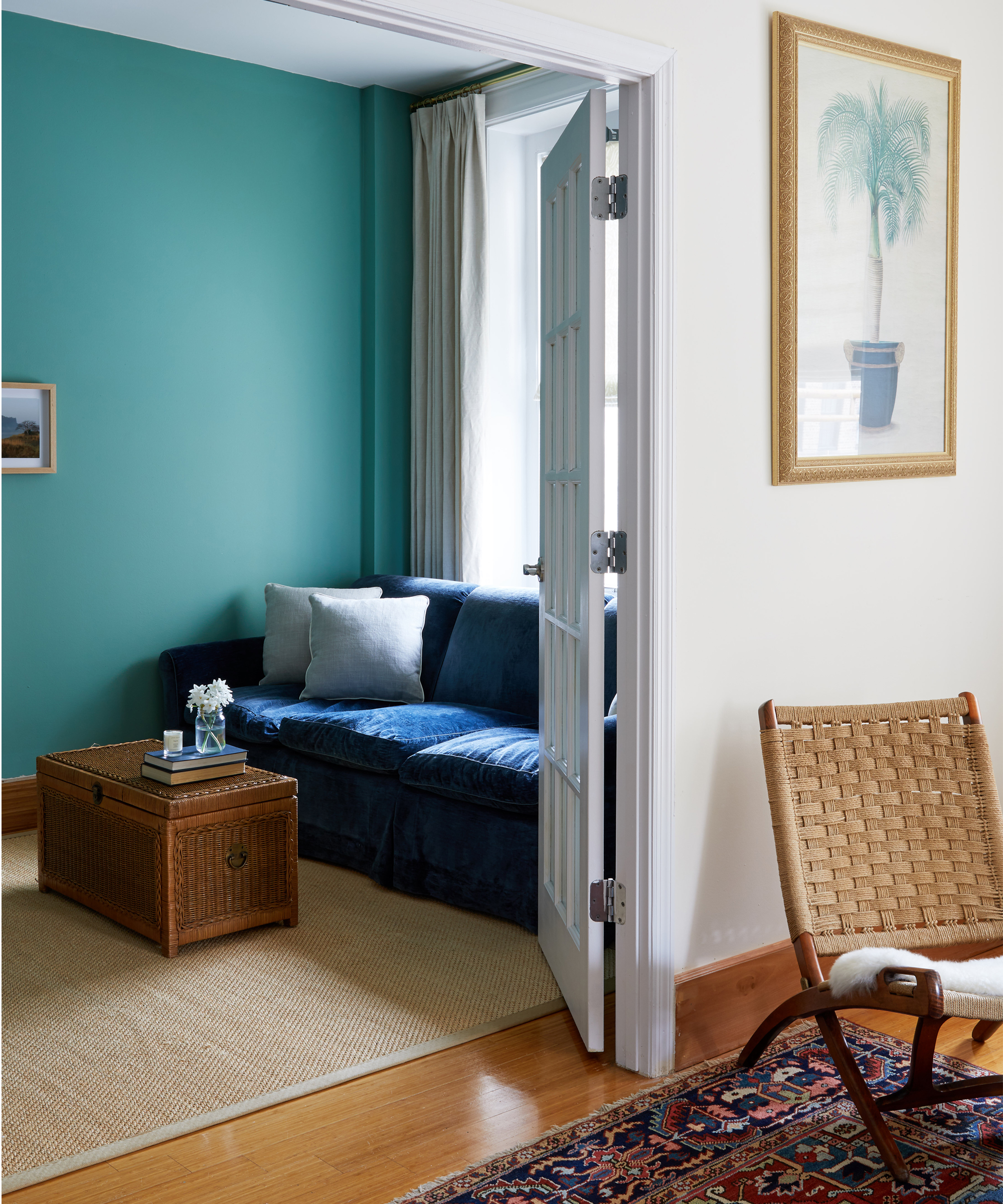 A cozy Davina Pike Wovn Home living room with a blue velvet sofa, wicker trunk coffee table, and cream rug. Teal and white walls feature large framed plant artwork, while sunlight pours in through an open door onto a woven chair.