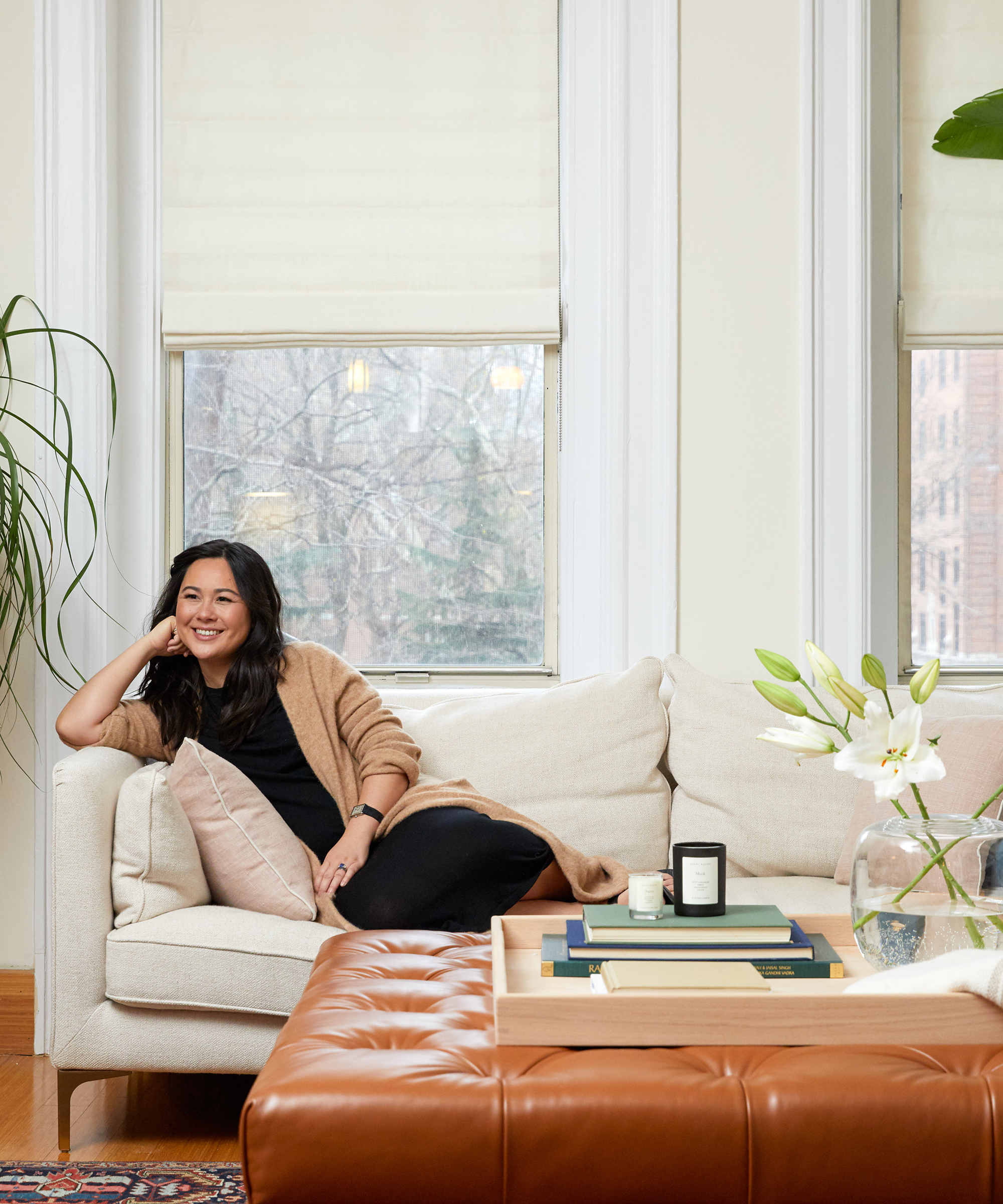 Davina Pike of Wovn Home sits smiling on a cream-colored sofa in a bright living room with large windows, a tan leather ottoman, and a vase of white flowers on a tray.