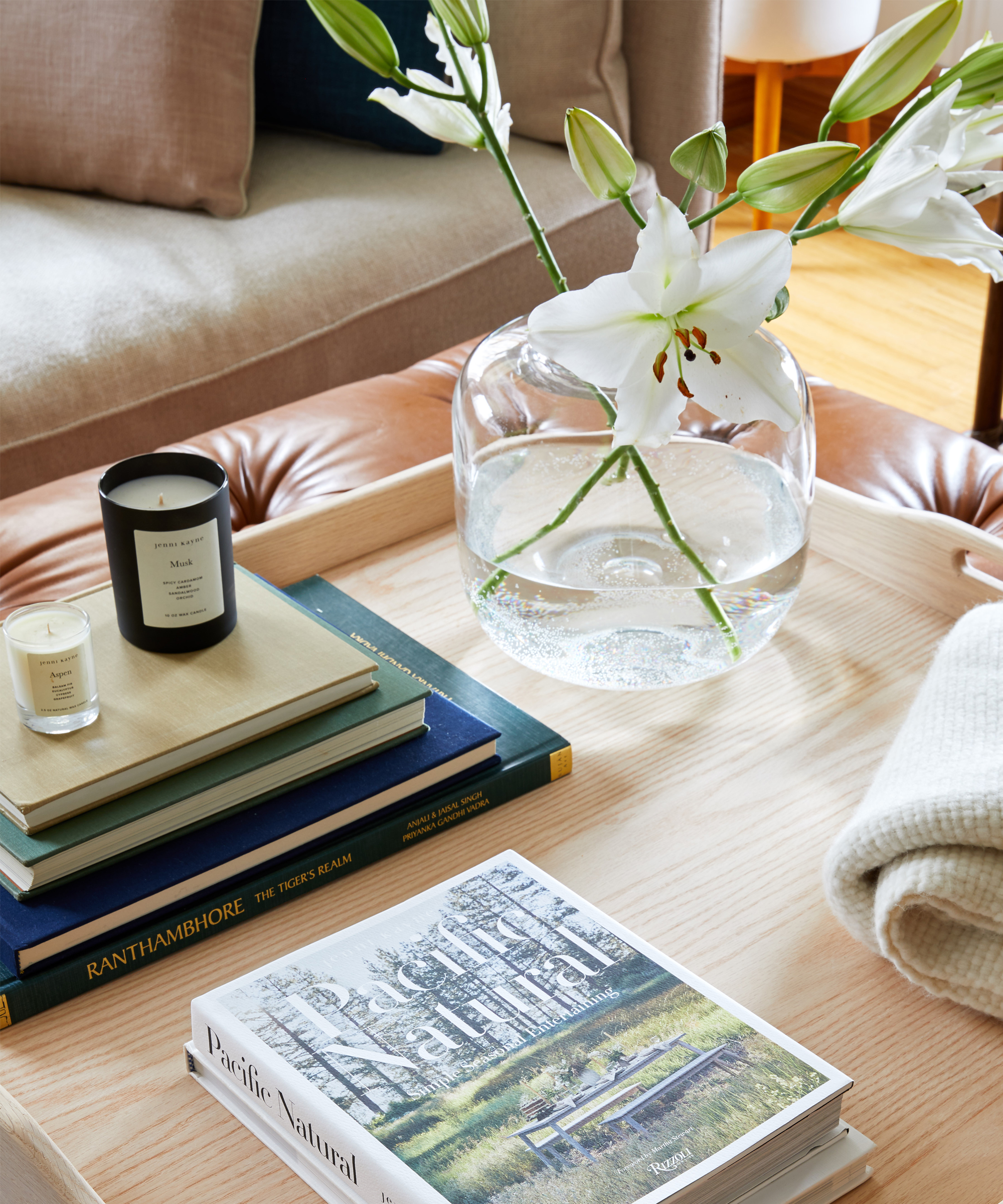 A close-up of a wooden coffee table with a glass vase holding white lilies, a stack of books, two candles, and a folded cream-colored blanket by Davina Pike for Wovn Home in a cozy, modern living room setting.