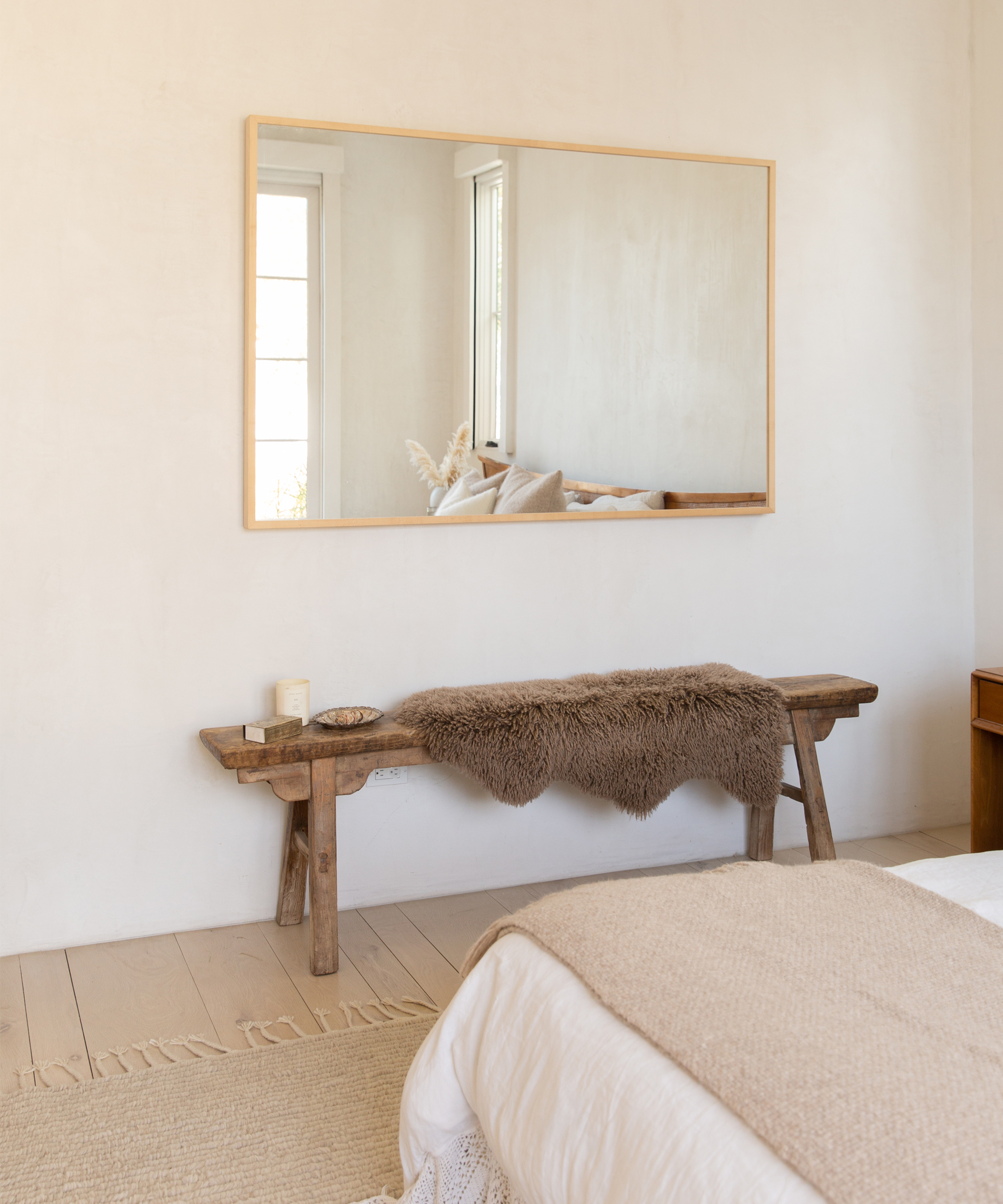 A minimalist bedroom features a wooden bench with a fur throw, a candle, and decor, placed below a large wall mirror. Inspired by Jenni Kayne Home, neutral tones and natural light create a calm, cozy atmosphere.