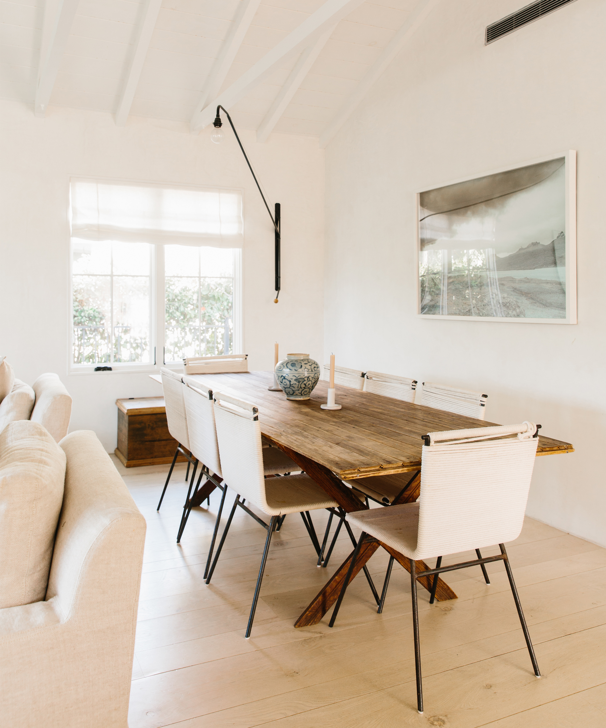 Bright dining room with a wooden table, six white chairs, and a decorative bowl, styled in the spirit of Julia Hunter and Jenni Kayne Home. Large window fills the space with light; neutral walls, floors, sofa, and framed art complete the look.