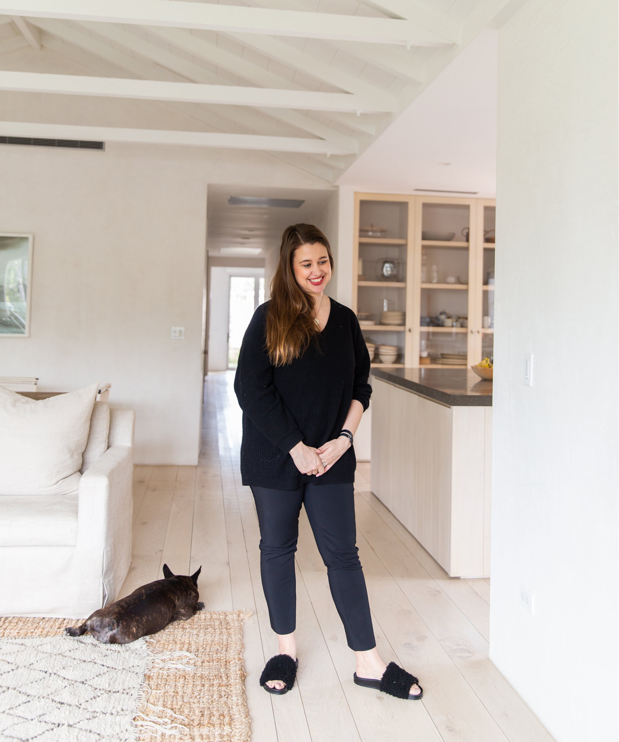 A woman with long brown hair, wearing a black sweater, black pants, and black slippers, stands smiling in a bright, modern Jenni Kayne Home living room. A small brown dog lies on the floor beside a beige sofa.