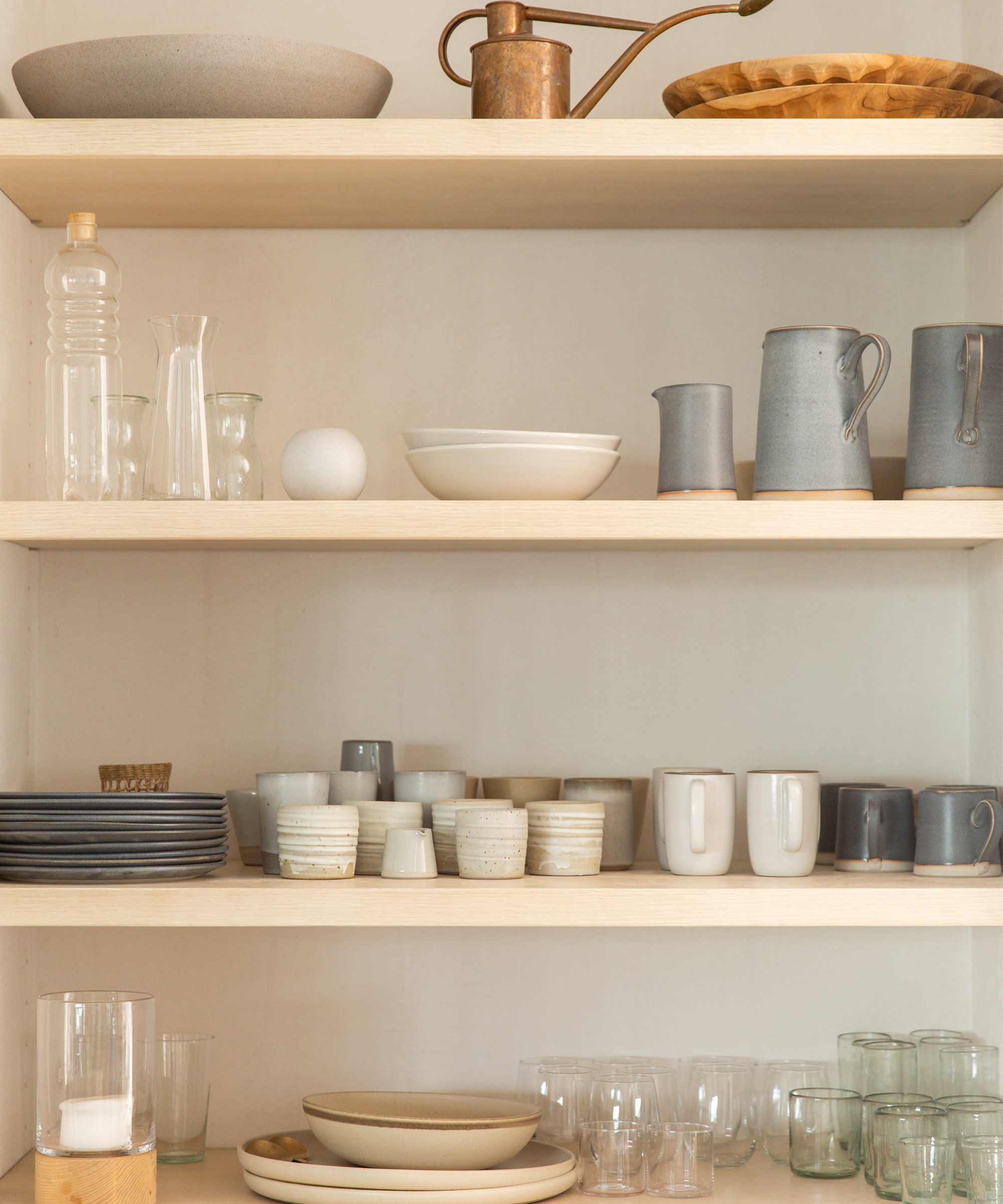 Open wooden shelves display neatly arranged ceramics, cups, plates, and glassware in soft neutral tones, echoing the calm elegance of a Julia Hunter Jenni Kayne Home-inspired minimalist kitchen.
