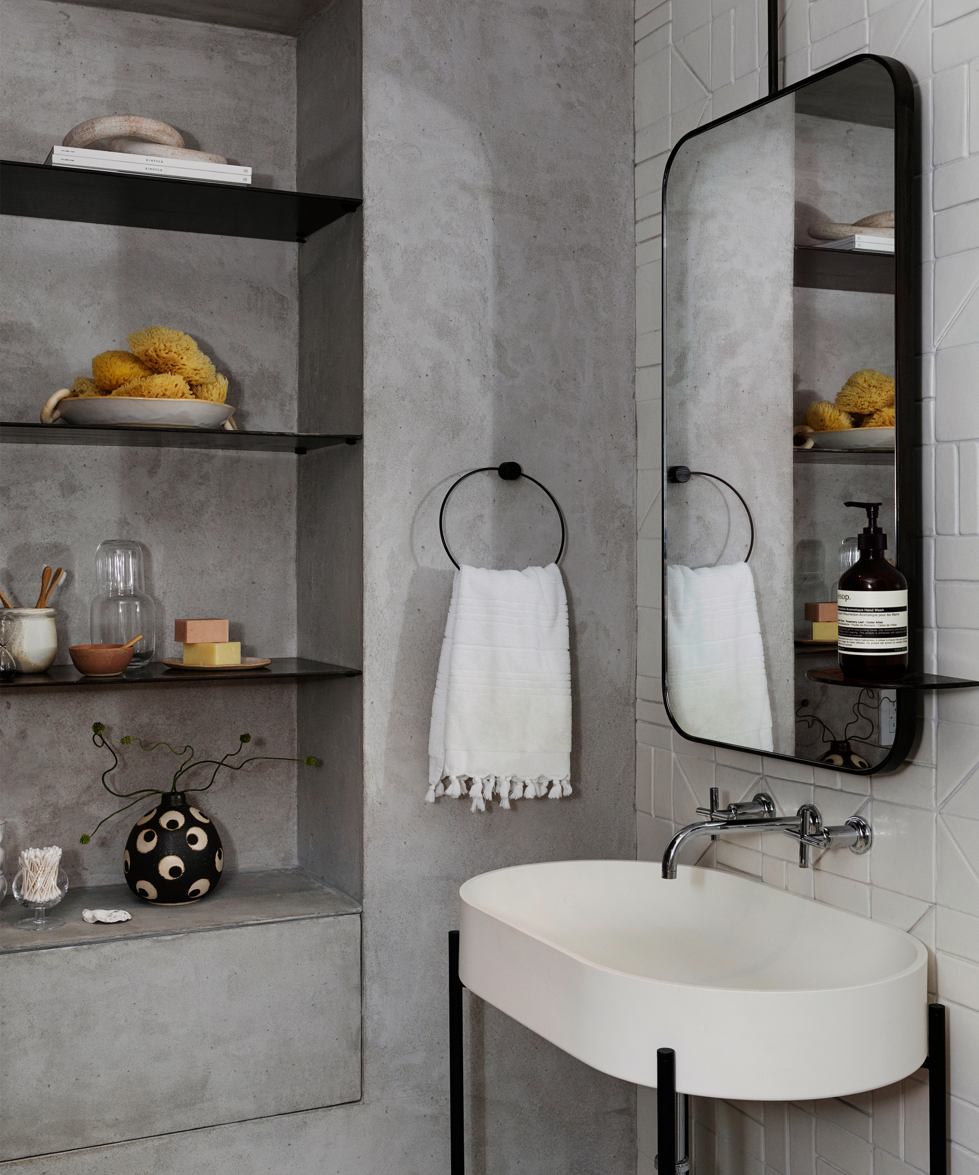 A modern bathroom inspired by tina rich interior design loft tour features concrete walls, open shelves with decor and towels, a rectangular wall-mounted mirror, a white sink with black metal legs, and a round towel ring holding a white hand towel.
