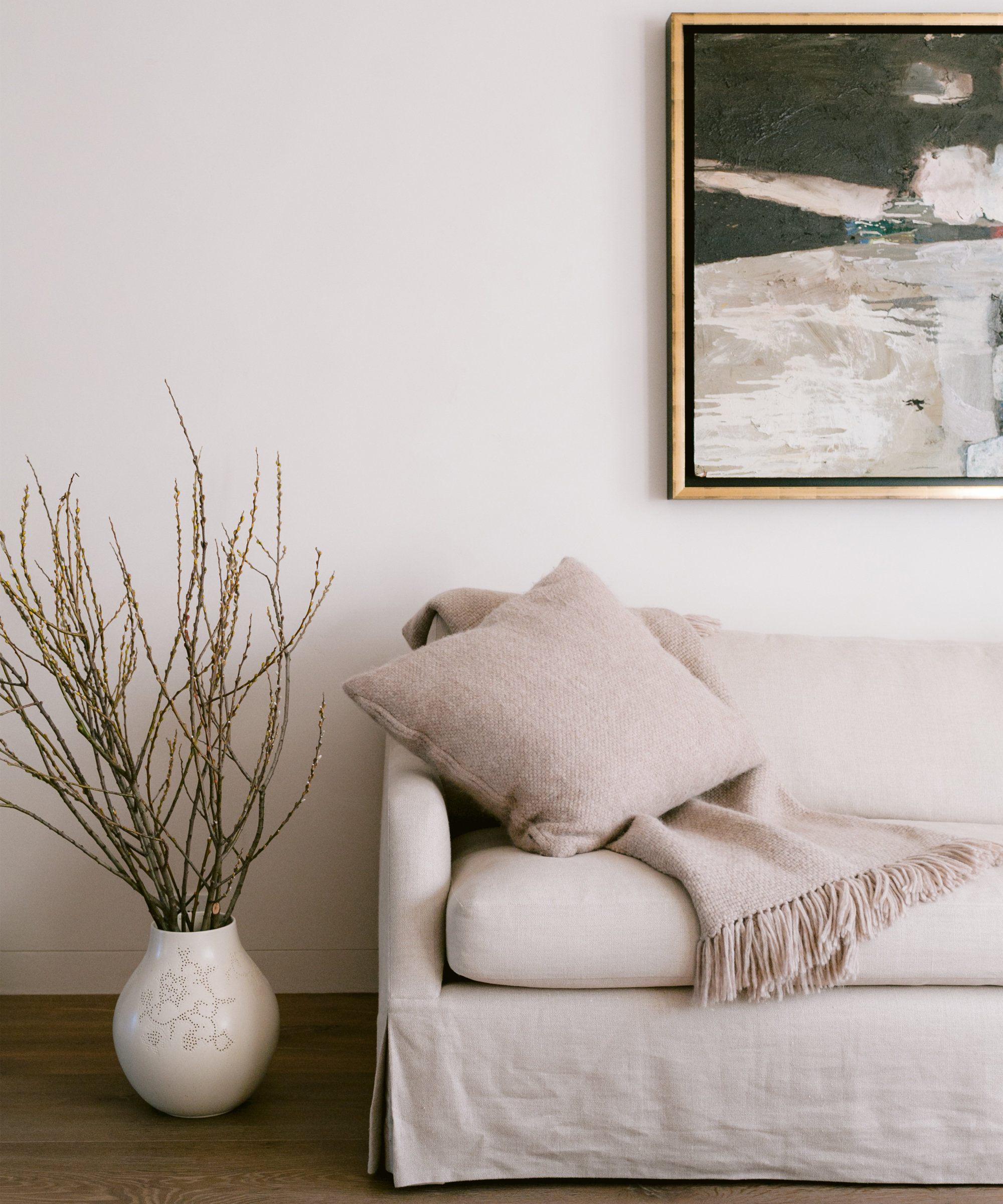 A beige sofa with a matching pillow and throw blanket sits near a white wall. A large vase with branches is on the floor, while an abstract painting inspired by April Gargiulo Vintner’s Daughter adds elegance to this calm, minimalist setting.