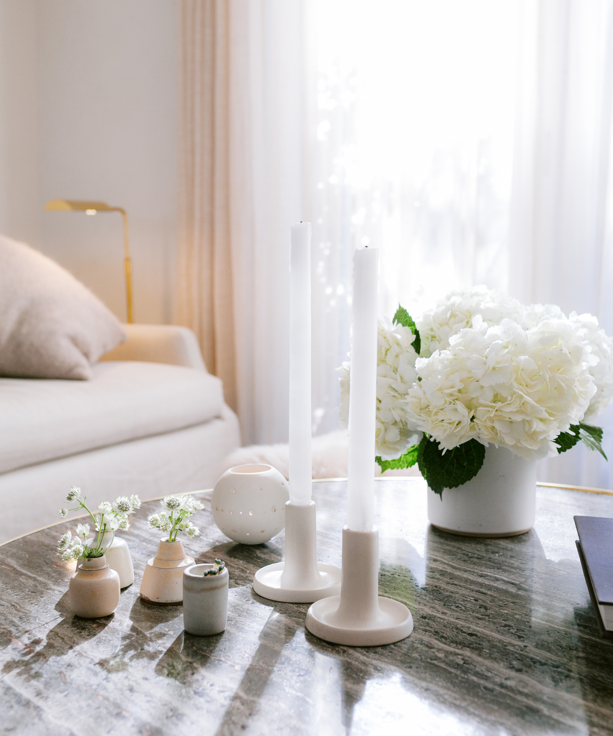 A marble coffee table displays two white candlesticks, small vases with tiny flowers, a white vase filled with hydrangeas, and a copy of April Gargiulo Vintner’s Daughter in a bright, cozy living room with a beige sofa and sheer curtains.