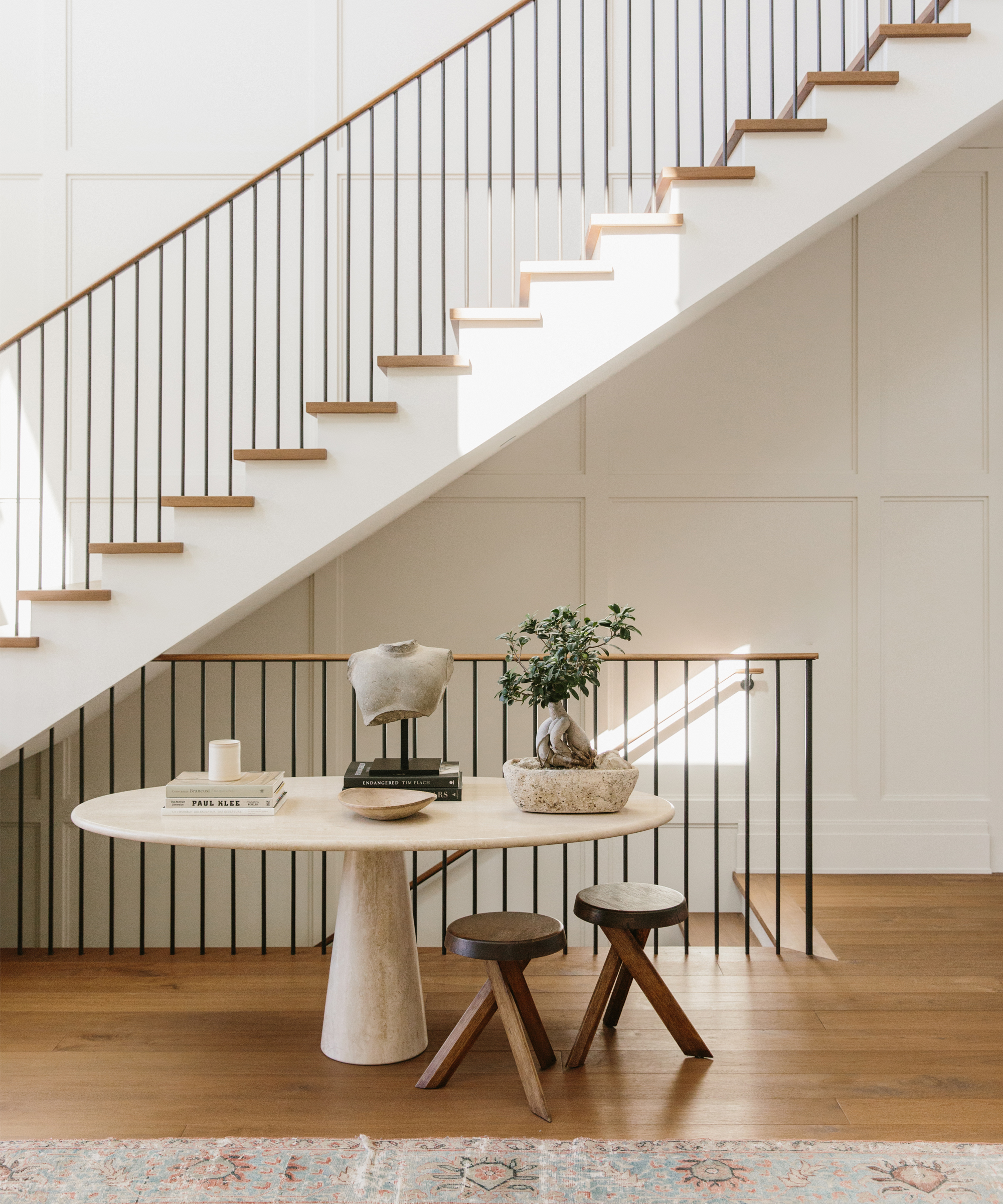 A minimalist entryway, inspired by Sheila Bouttier Galerie Provenance Home Tour, features a round table with decor items, two wooden stools, and a potted plant beneath a modern staircase. Light streams in, highlighting the clean, neutral design.