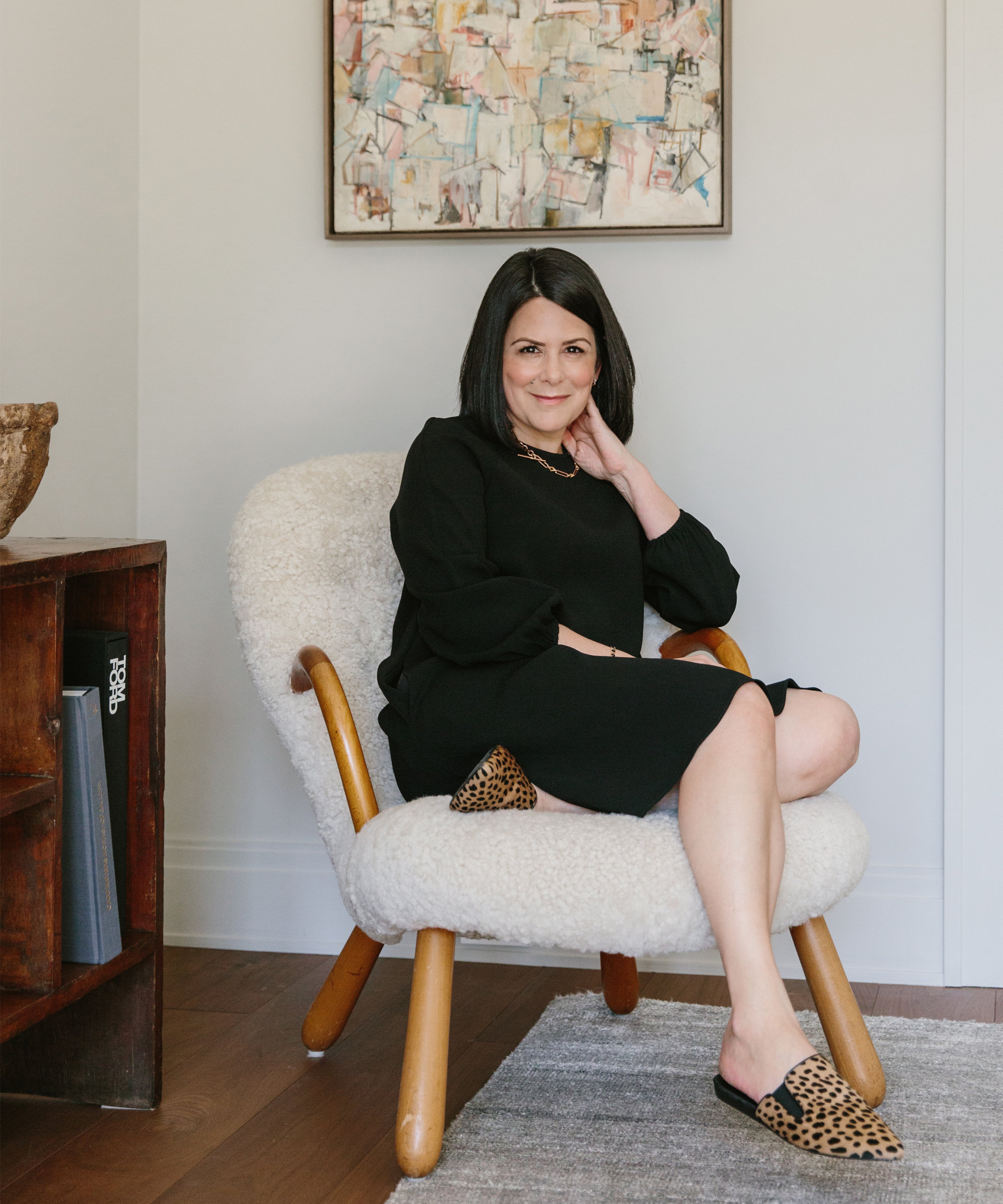 Sheila Bouttier sits relaxed in a cozy white armchair, wearing a black dress and leopard-print shoes. She smiles warmly, her head resting on her hand, with abstract art from Galerie Provenance displayed behind her during a serene home tour.