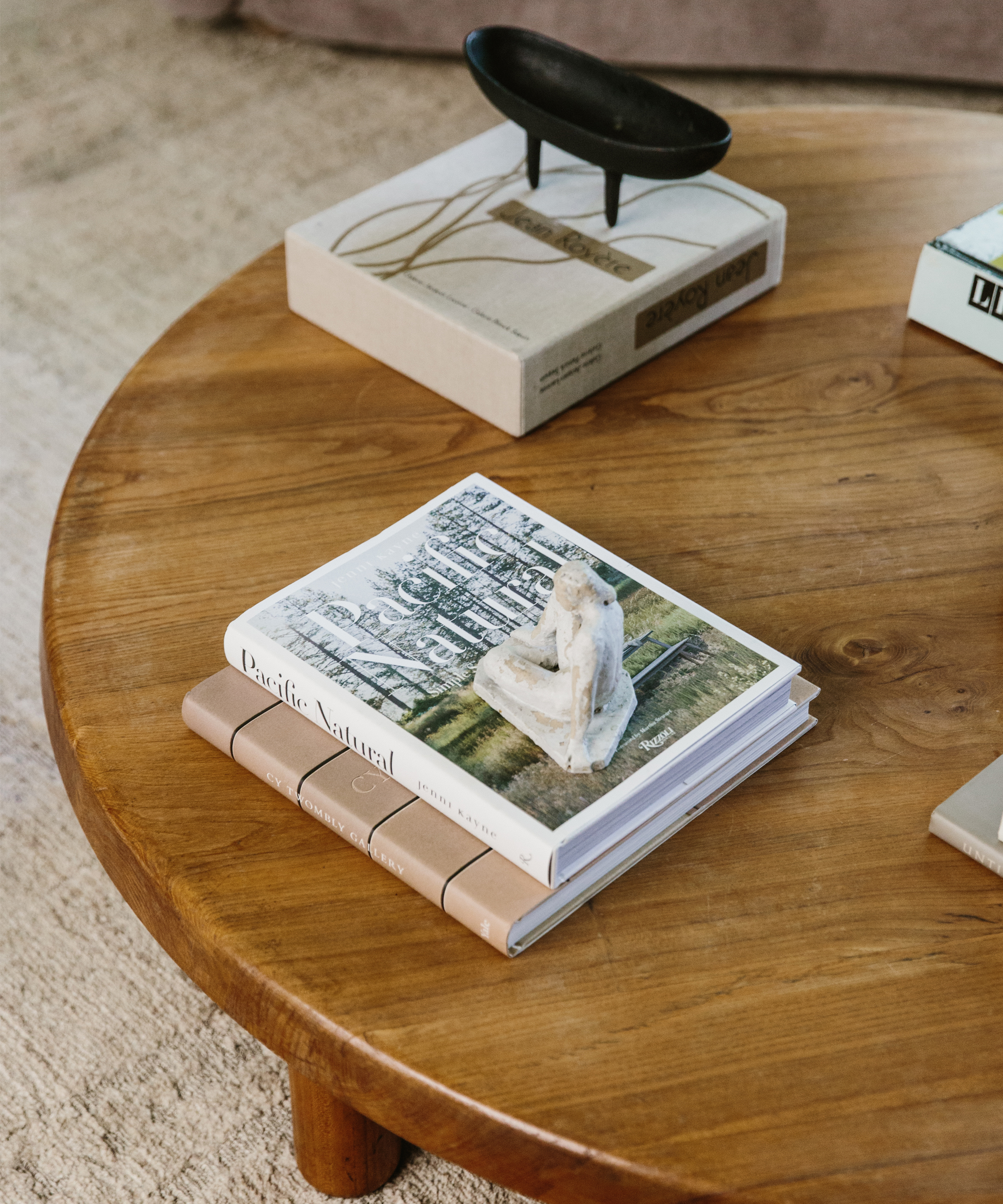 A round wooden coffee table, as seen in the Sheila Bouttier Galerie Provenance Home Tour, displays stacked books—one topped with a small white statue and another with a decorative black dish—all resting on a textured beige rug.