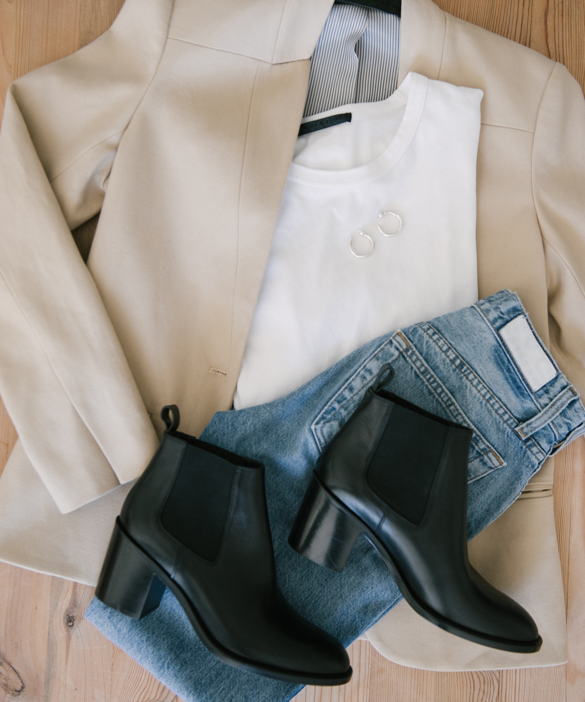 A beige blazer, white T-shirt, silver hoop earrings, light blue jeans, and black heeled ankle boots are neatly arranged on a wooden surface—perfect inspiration for chic blazer outfits.