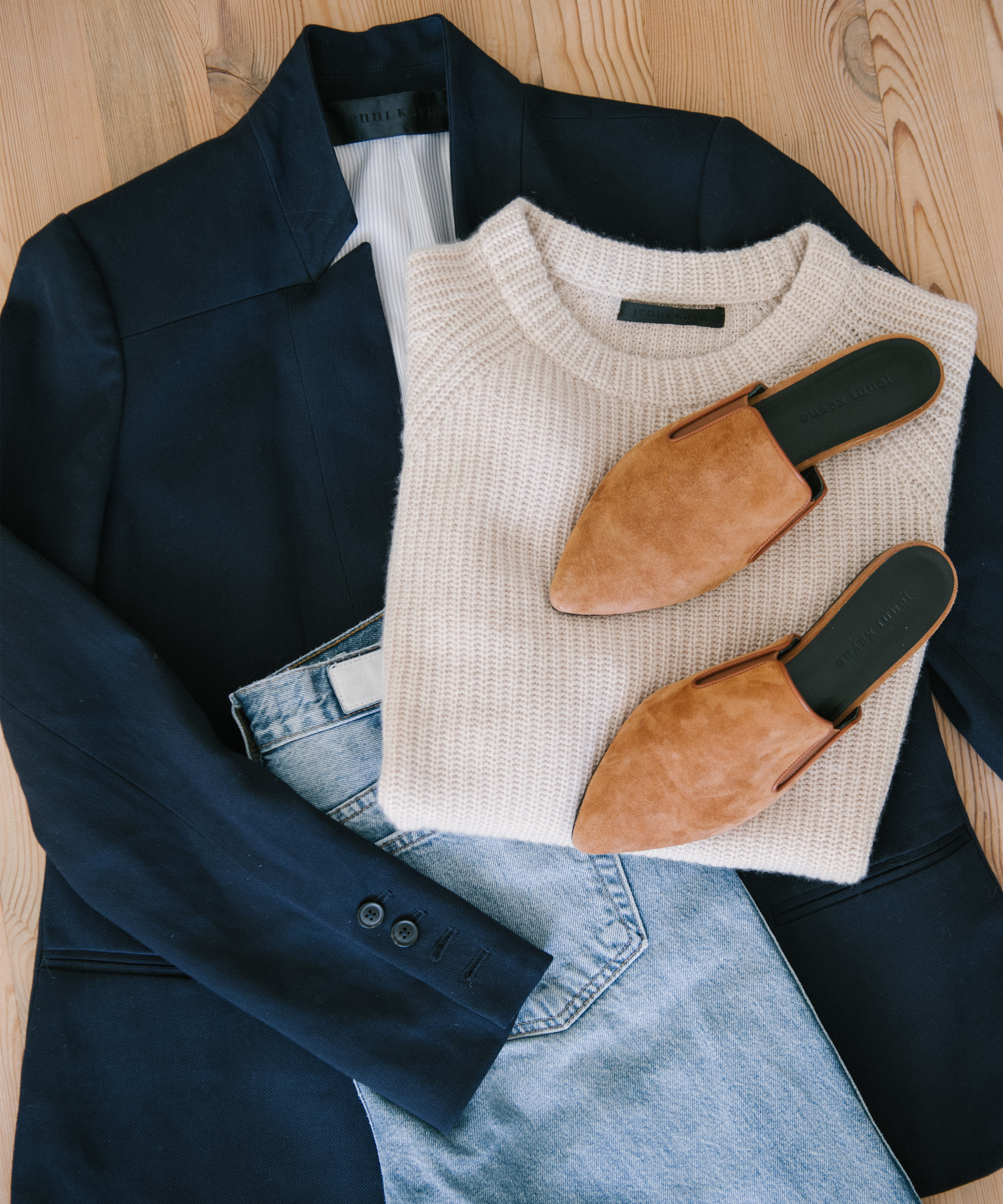 Flat lay of a chic blazer outfit featuring a navy blazer, cream knit sweater, light blue jeans, and tan suede pointed-toe mules on a wooden surface.