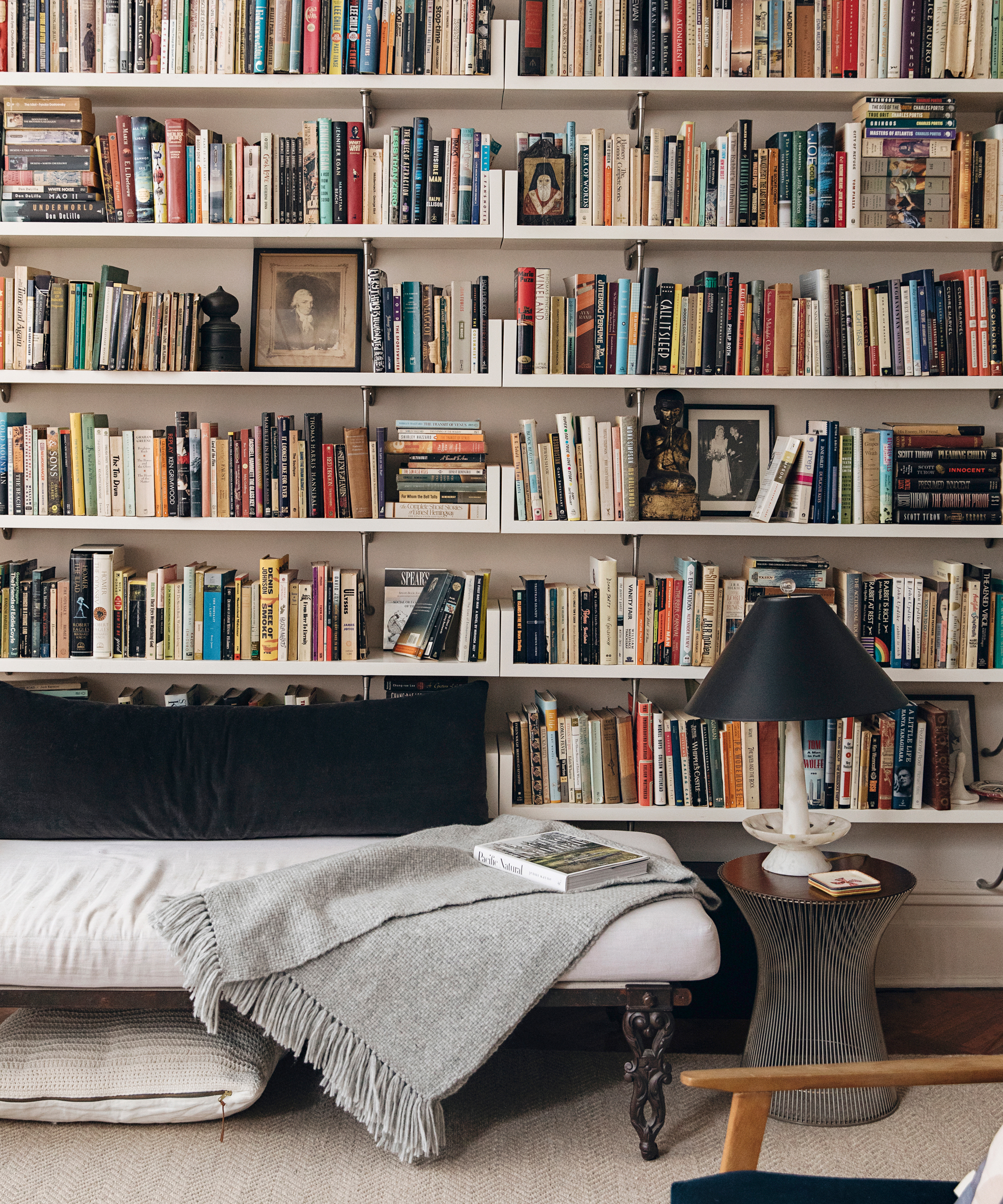 A cozy reading nook with a cushioned bench, a gray throw blanket, a side table with a lamp, and tall shelves filled with books—including Amanda Hesser’s favorites from Food52—framed photos, and small decorative objects.
