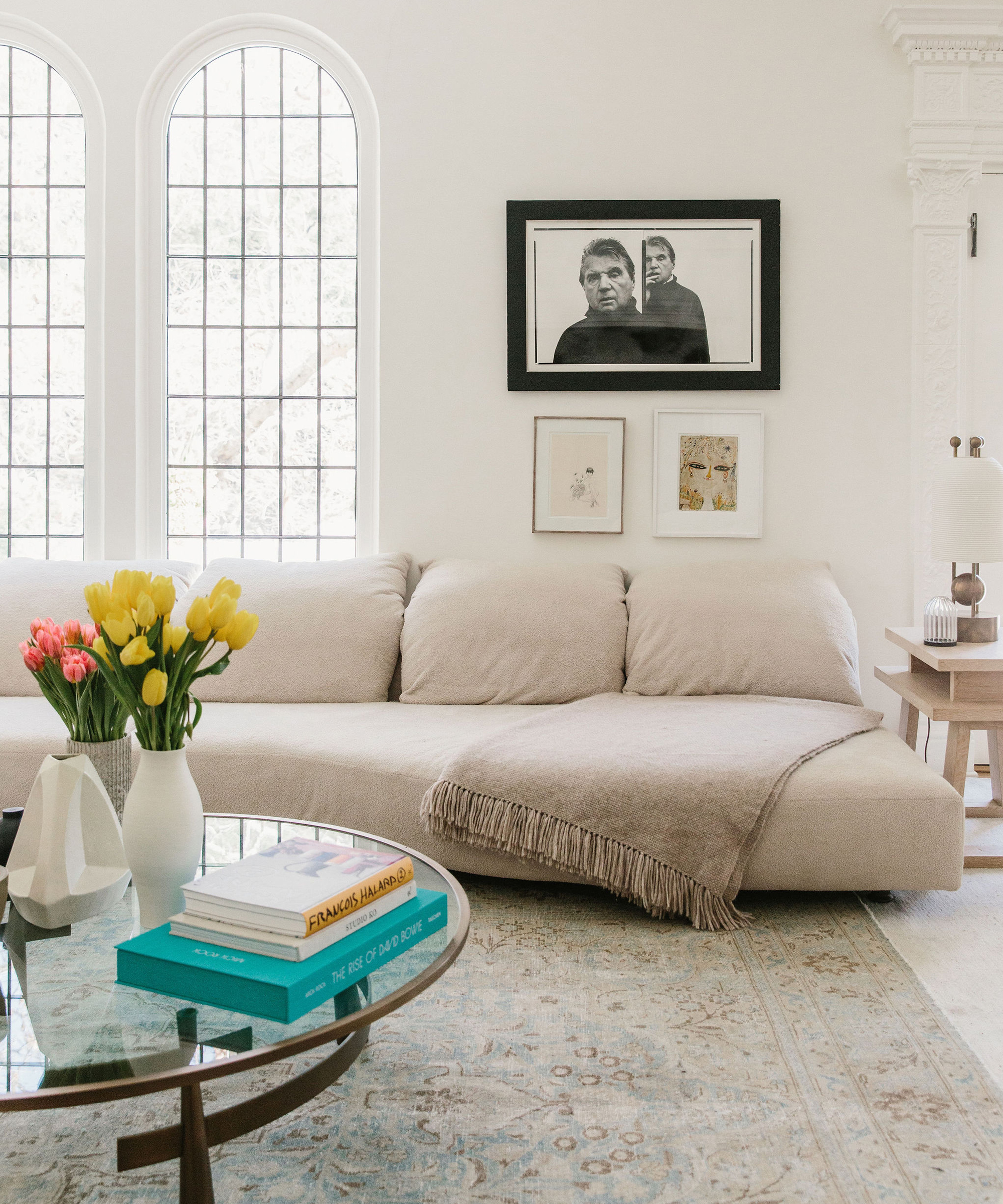 Table with coffee table books and tulips in front of large oatmeal colored couch in Brigette Romanek's living room under black and white framed photograph