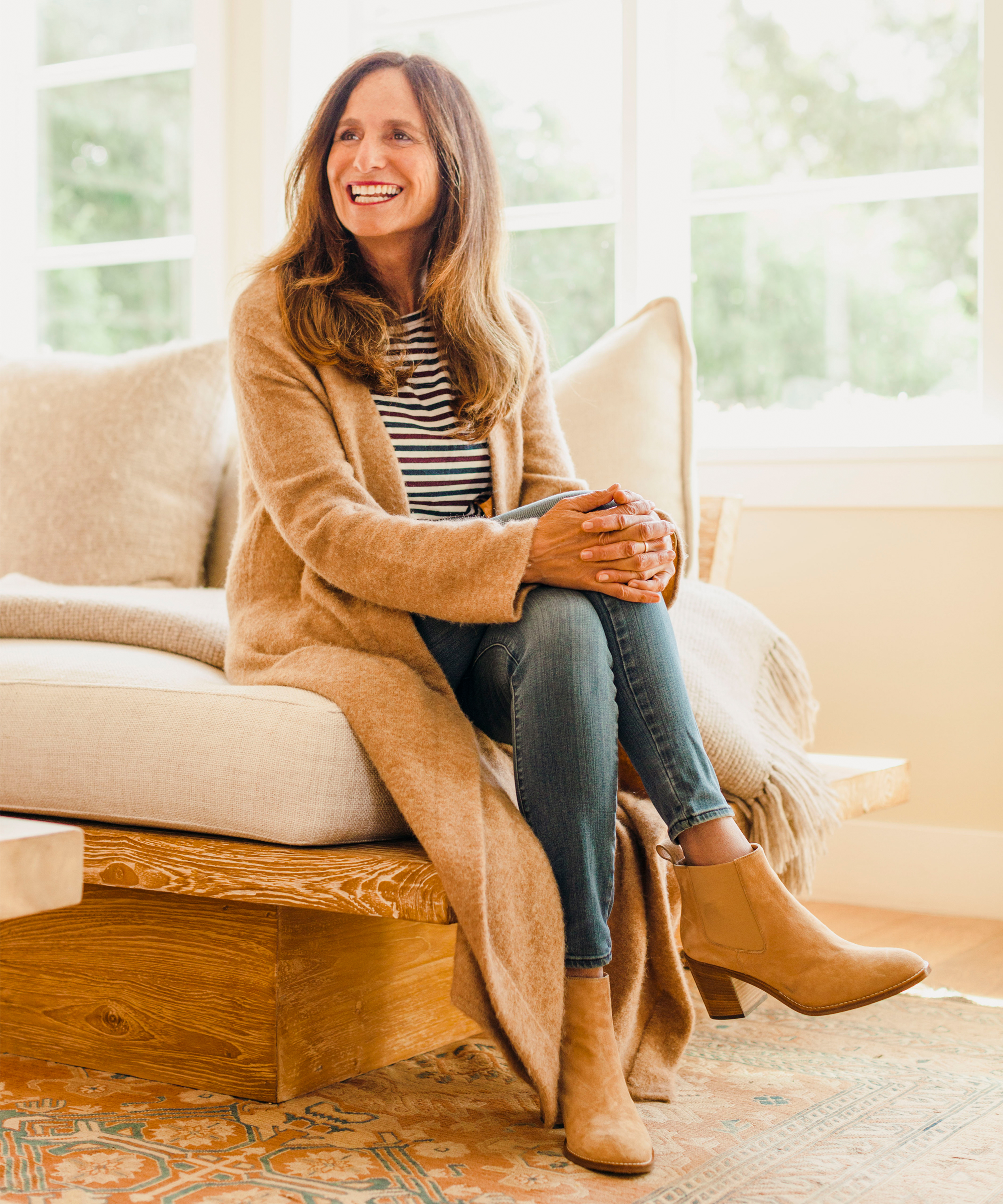 A woman with long brown hair sits on a wooden couch, smiling as she reads her astrology birth chart. She wears a tan coat, striped shirt, blue jeans, and tan ankle boots in a bright room with large windows and cozy, neutral-toned decor.