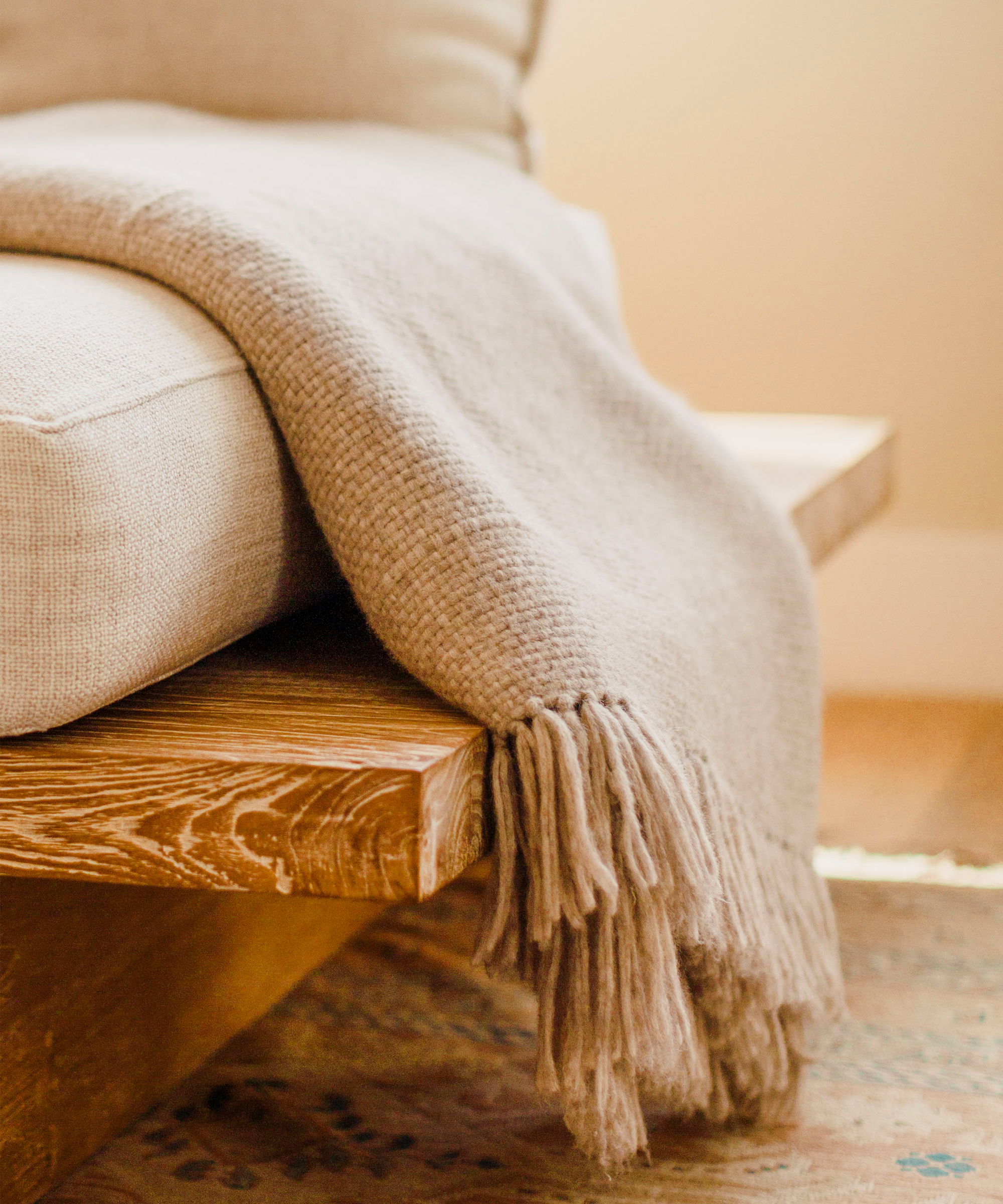 A beige, fringed throw blanket draped over the arm of a wooden sofa with light-colored cushions, set on a patterned rug in a cozy, softly lit room perfect for reading or exploring your astrology birth chart.