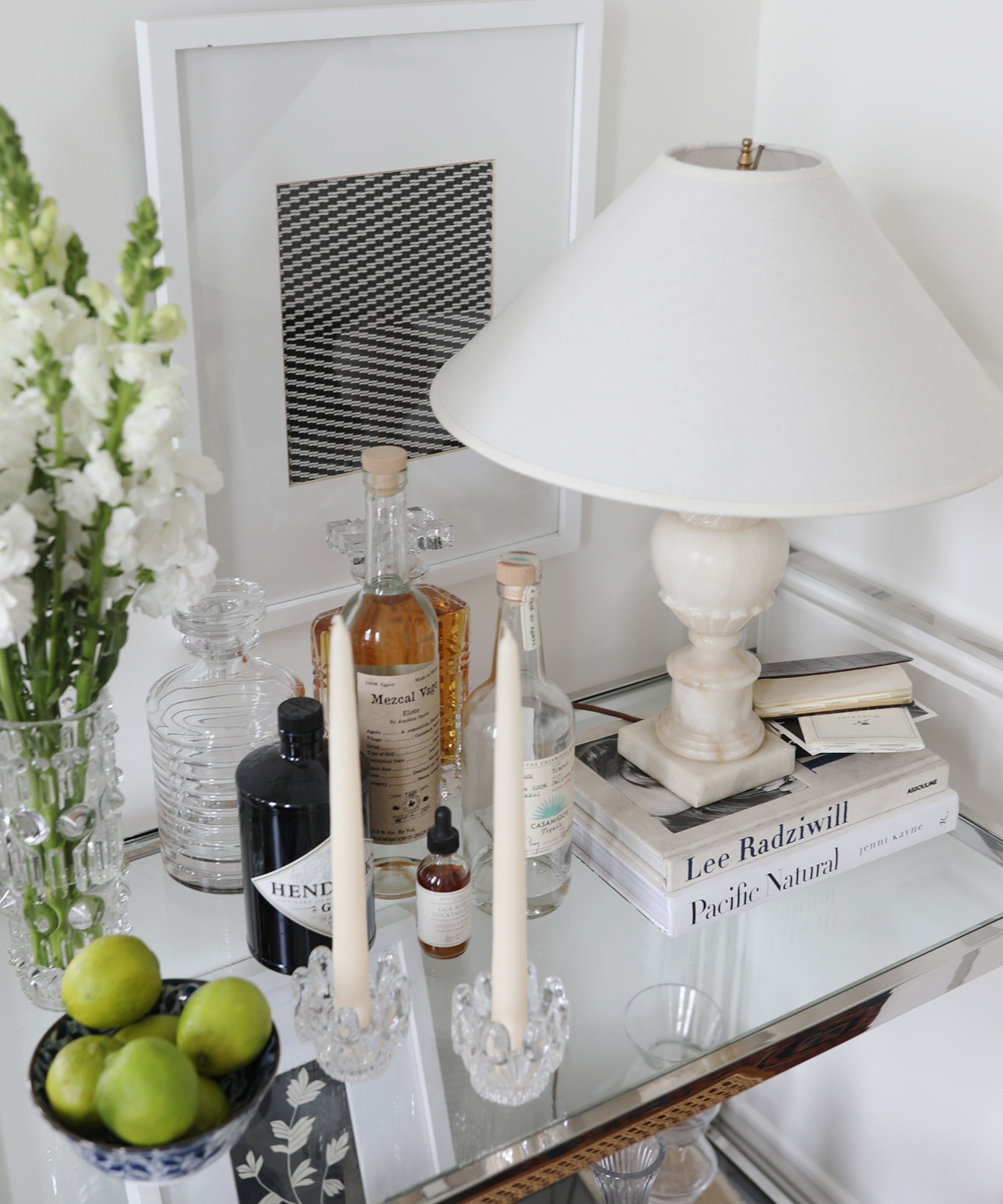 A mirrored bar cart styled by Ashley Kane holds bottles, a lamp, candles, books, a crystal vase with white flowers, a bowl of limes, and framed art against a white wall for an elegant, curated look.