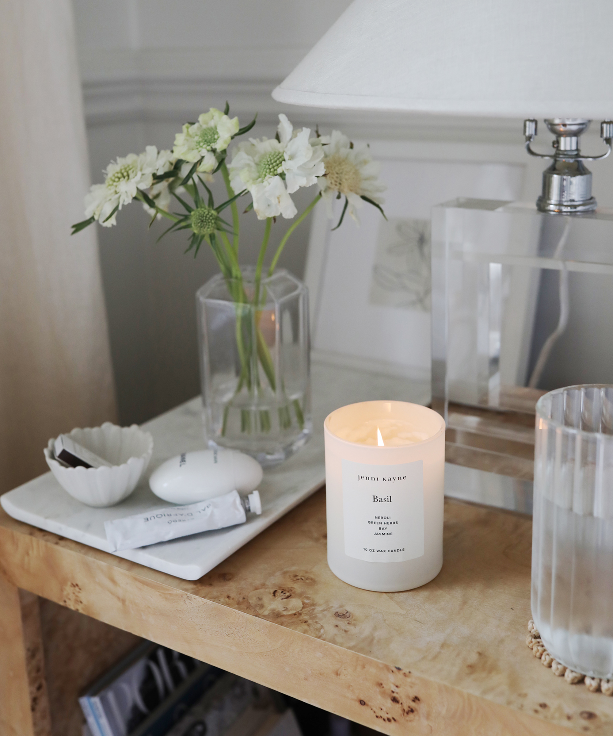 A lit white candle sits on a wooden bedside table next to a glass of water, a clear vase with white flowers, and a small white dish in this serene Ashley Kane-inspired setting. A lamp with a white shade glows softly in the background.