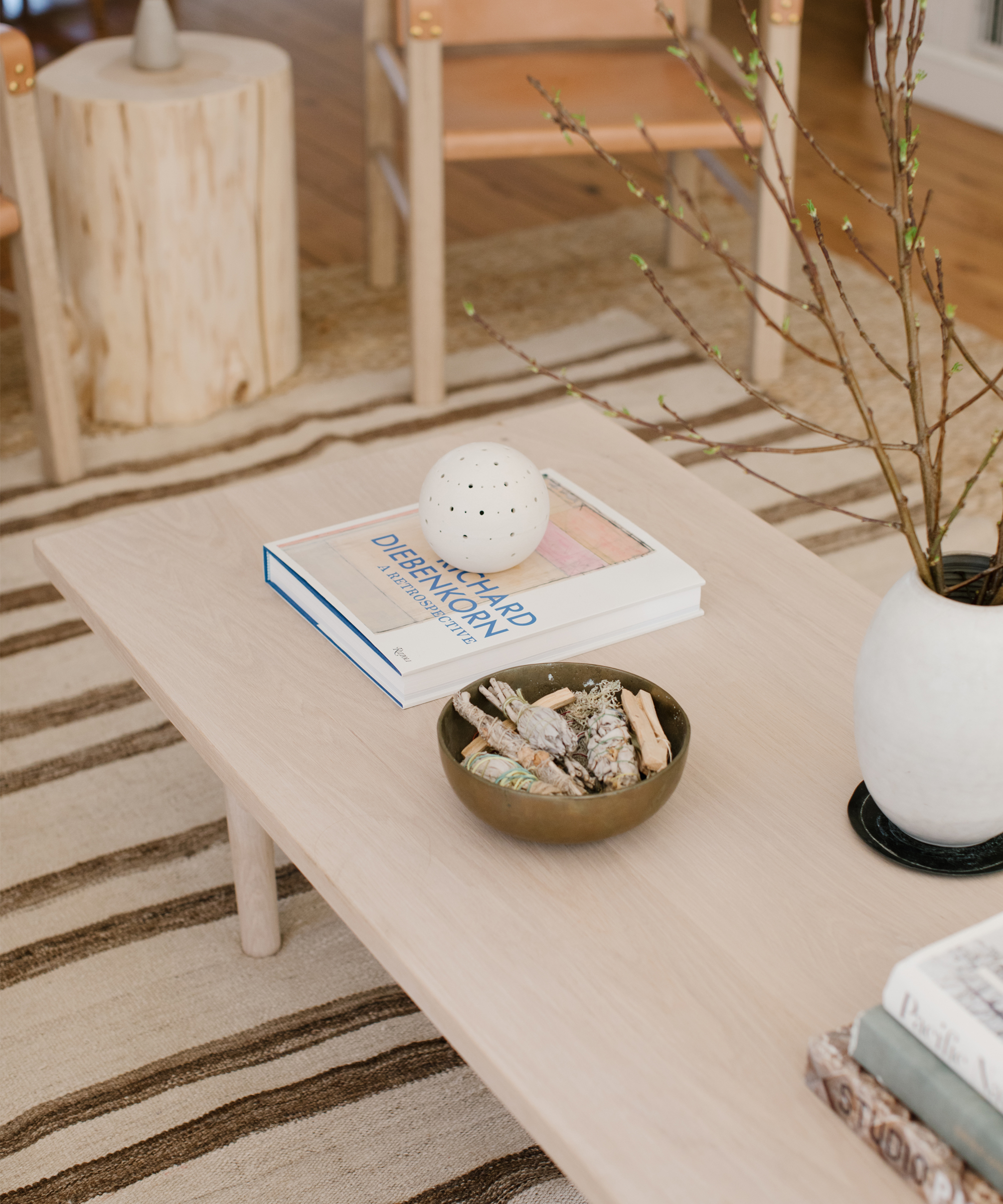 A light wooden coffee table demonstrates how to style your coffee table with a bowl of decorative objects, books, a round white ornament, and a white vase with branches, all set on a striped rug in a cozy living room.