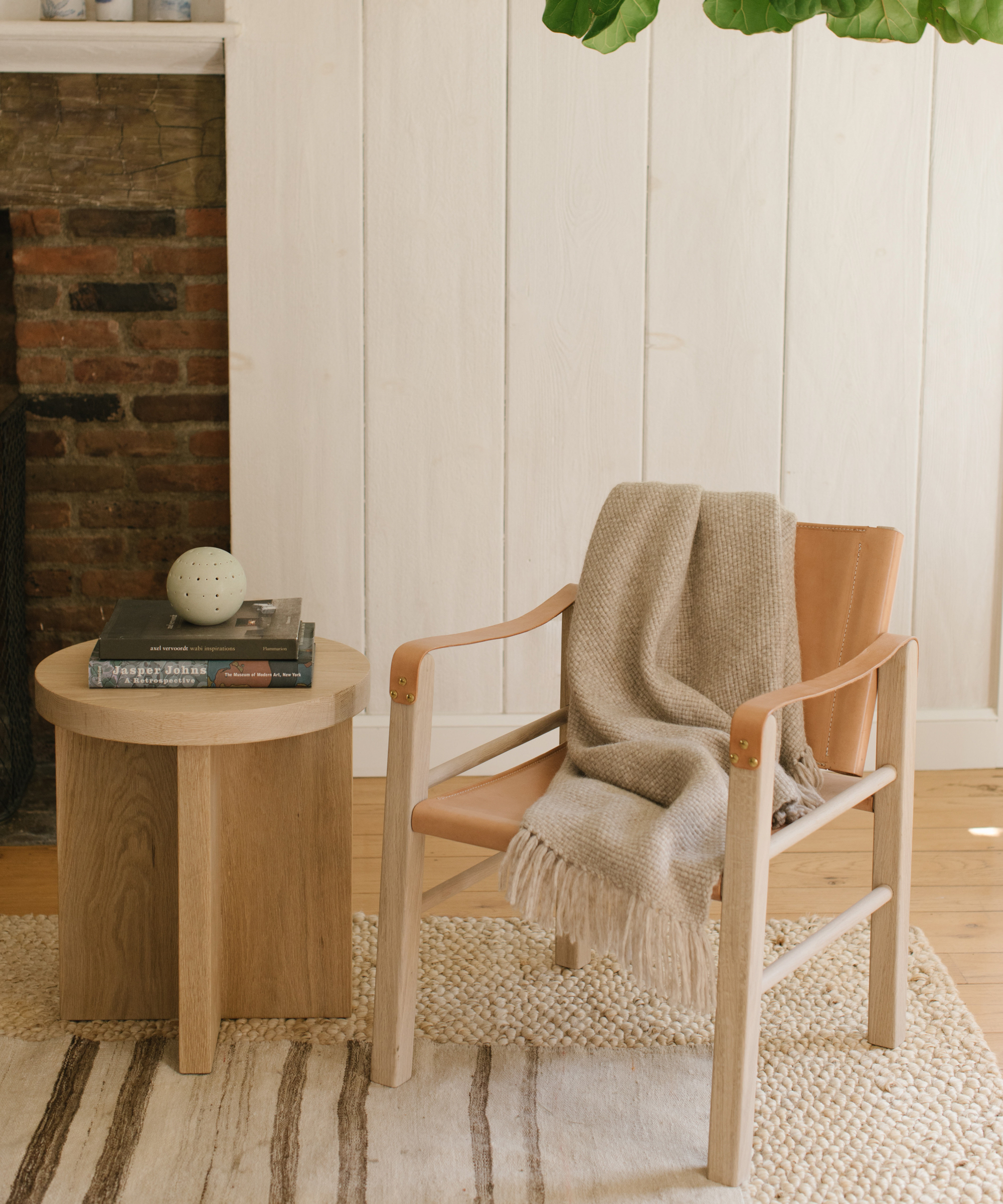 A tan leather armchair with a beige knit blanket draped over one arm sits beside a round wooden side table—offering cozy living room design ideas on a neutral rug near a white paneled wall and brick fireplace.