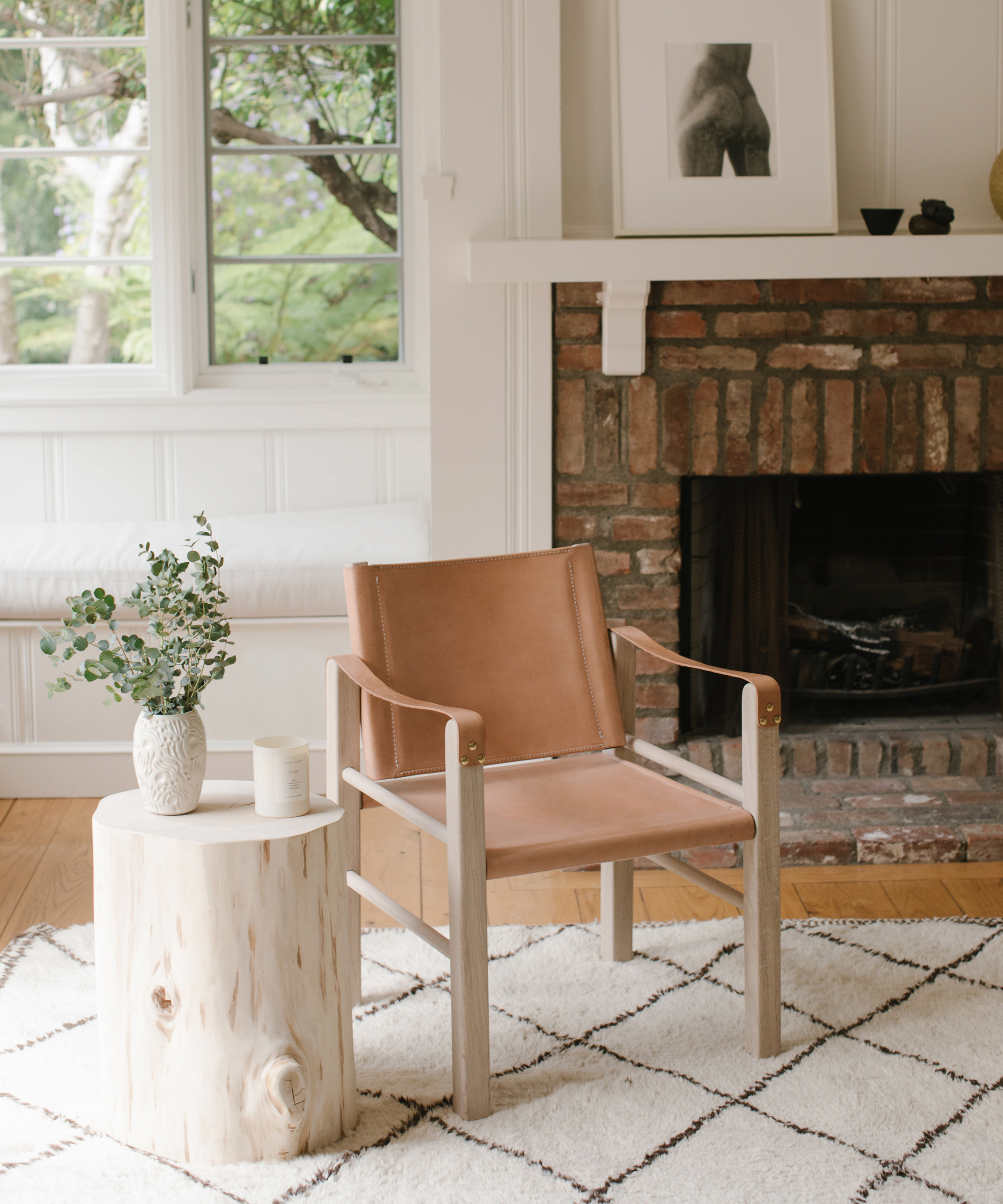 A light brown leather chair sits on a white rug with a diamond pattern, next to a round wooden side table with a vase of greenery and a candle—perfect inspiration for those seeking stylish living room design ideas. A brick fireplace and large windows are in the background.