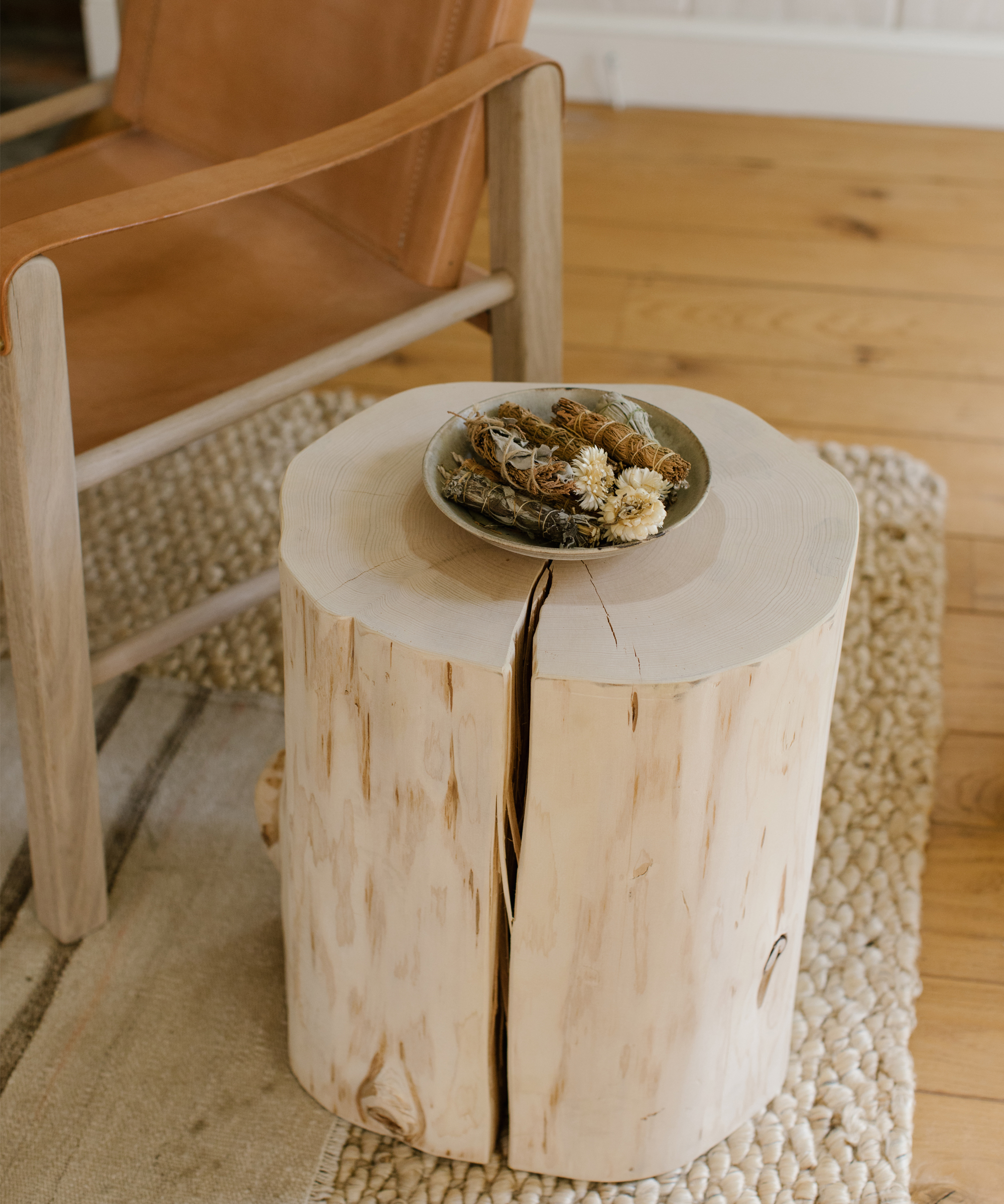 A rustic wooden stump table with a bowl of dried flowers, next to a tan leather chair, sits on a textured woven rug—an inviting touch for those seeking unique living room design ideas.