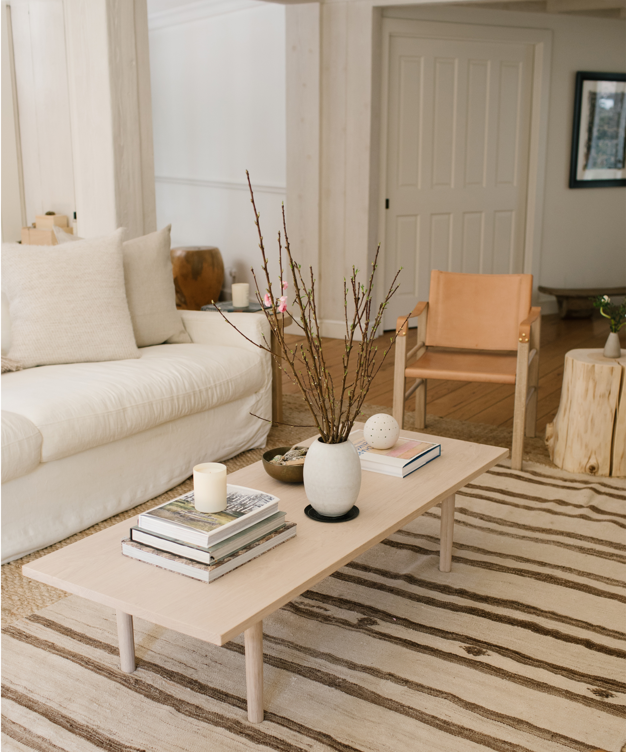 A minimalist living room with a white sofa, light wood coffee table holding books, a vase with branches, and a candle. A tan armchair and striped rug on wooden floors complete this space—perfect for fresh living room design ideas.