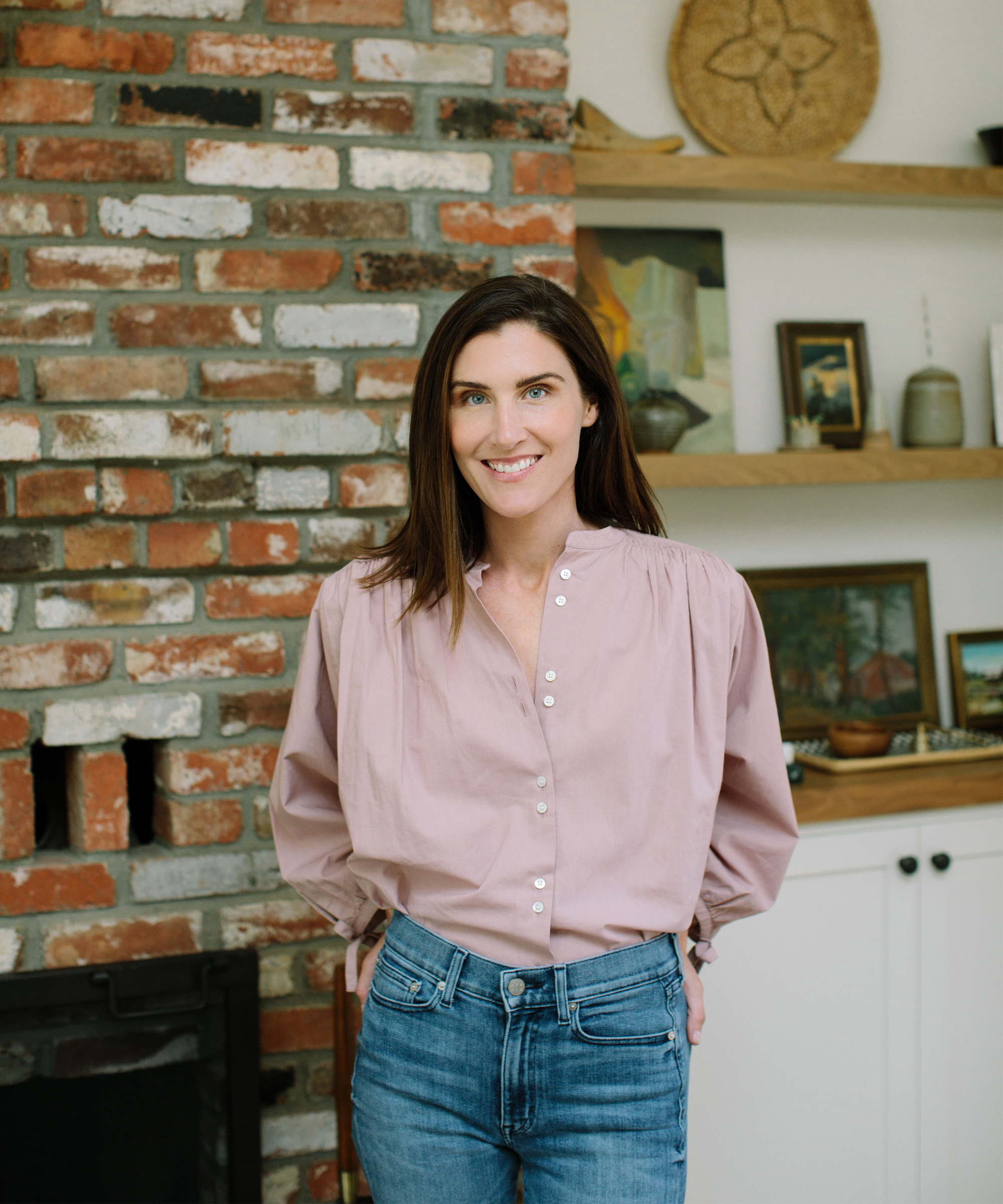 A woman with long brown hair, wearing a light pink button-up blouse and blue jeans, stands smiling indoors in front of a brick fireplace and wooden shelves decorated with art, pottery, and reminders of mindfulness practices.