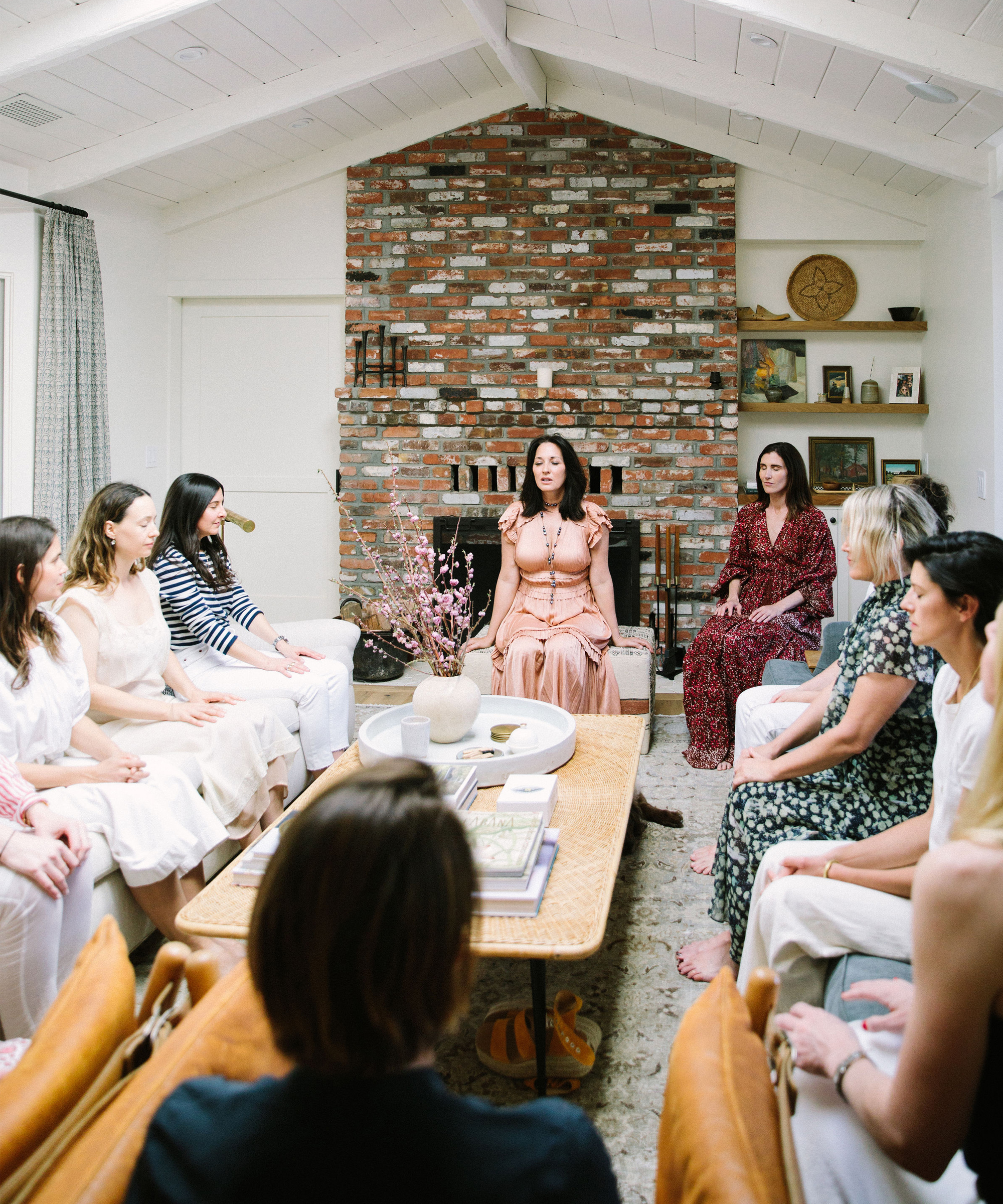 A group of women sits in a circle in a cozy living room with a brick fireplace, attentively listening to a woman in a pink dress at the center as she shares mindfulness practices.