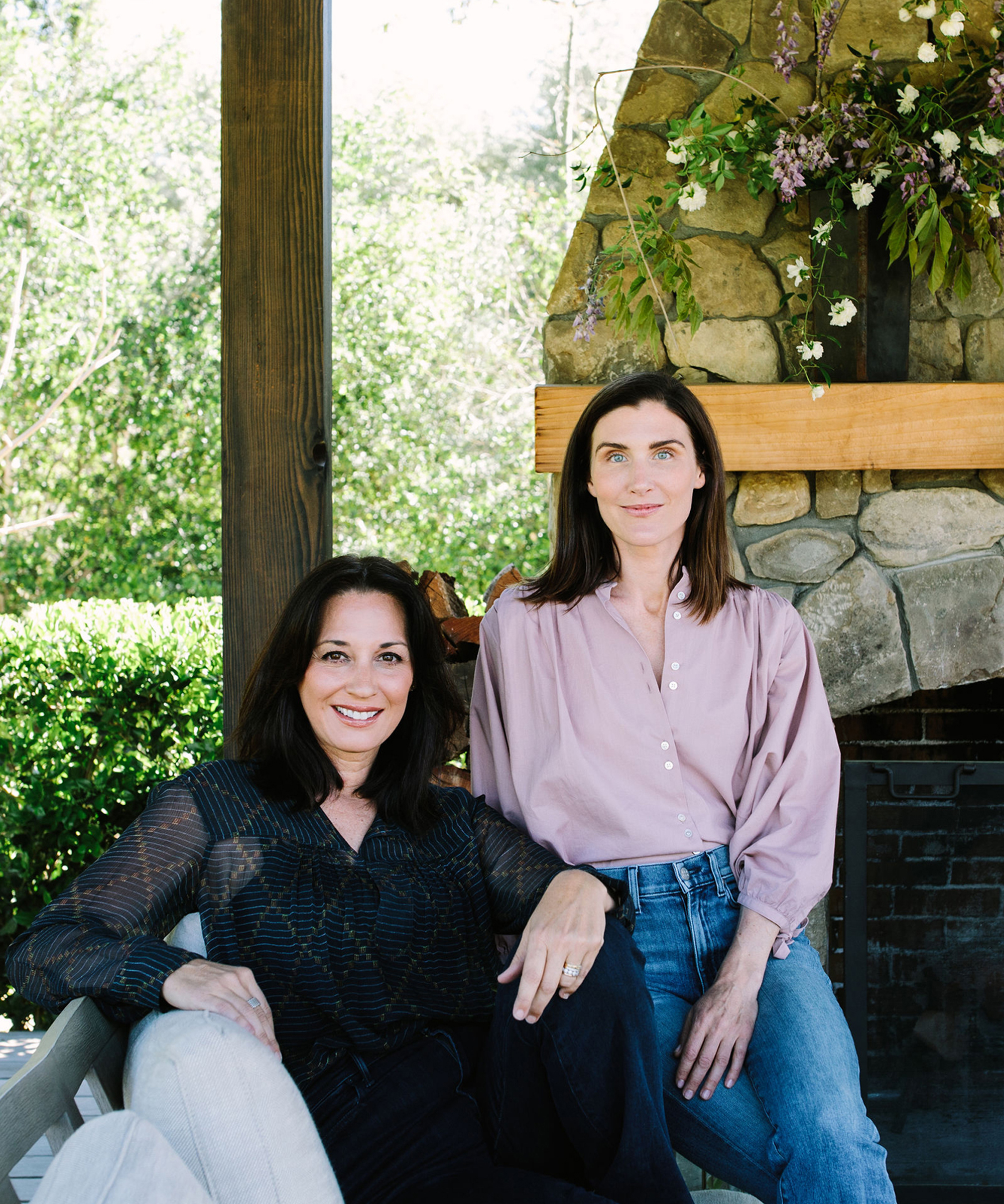 Two women pose together outdoors in front of a stone fireplace decorated with flowers. Their serene smiles and relaxed postures evoke the essence of mindfulness practices, with lush greenery providing a peaceful background.